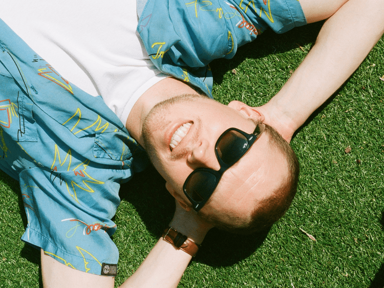Young man lying on grass, smiling, wearing sunglasses, a patterned blue shirt, and a wristwatch.