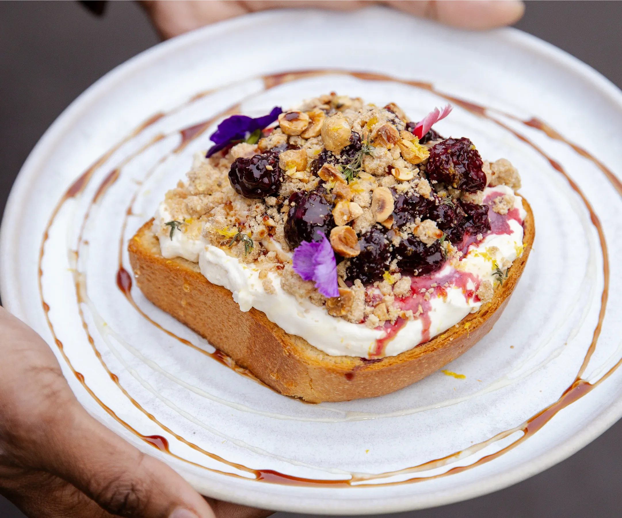 Close-up of a dessert with whipped cream, berry compote, nuts, edible flowers, and honey syrup on a white plate.