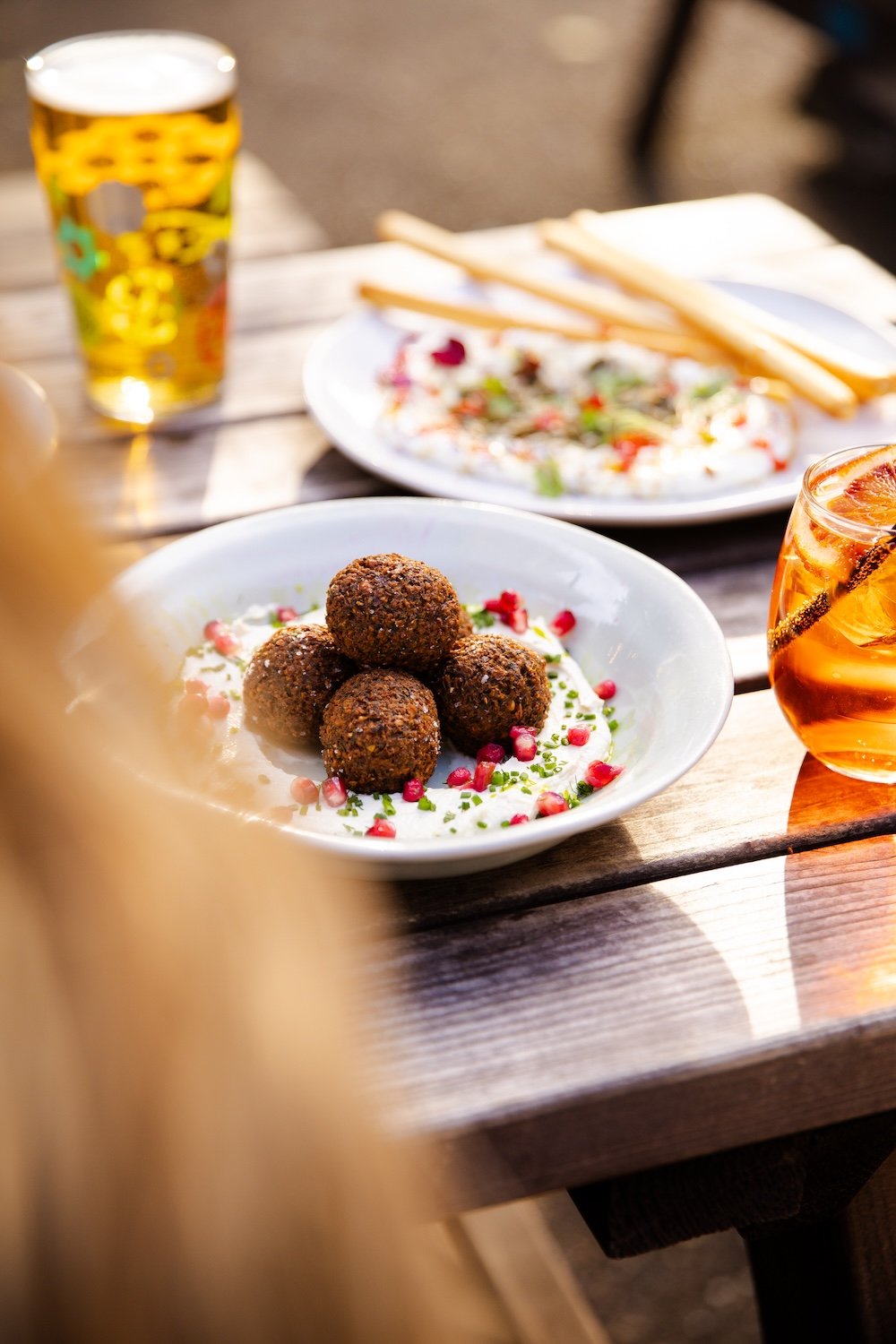 A plate of falafel balls garnished with pomegranate seeds and herbs, with a glass of beer and a soda on a wooden outdoor table.