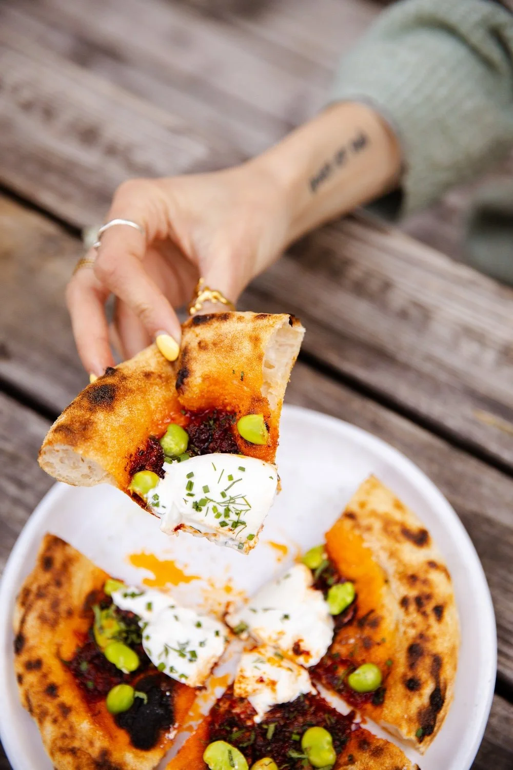 Person holding a slice of pizza with pesto, cheese, and toppings. The pizza is on a white plate on a wooden table.