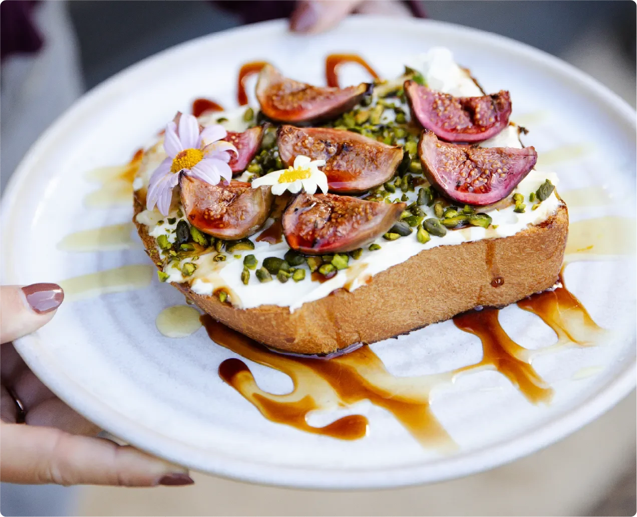 A slice of sweet bread topped with yogurt, sliced figs, chopped pistachios, edible flowers, honey, and caramel drizzle.