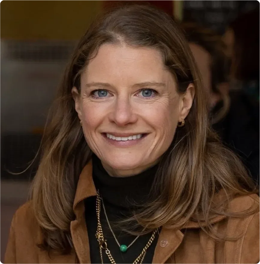 A woman with shoulder-length brown hair and blue eyes, smiling, wearing a brown jacket, black turtleneck, and layered gold necklaces.