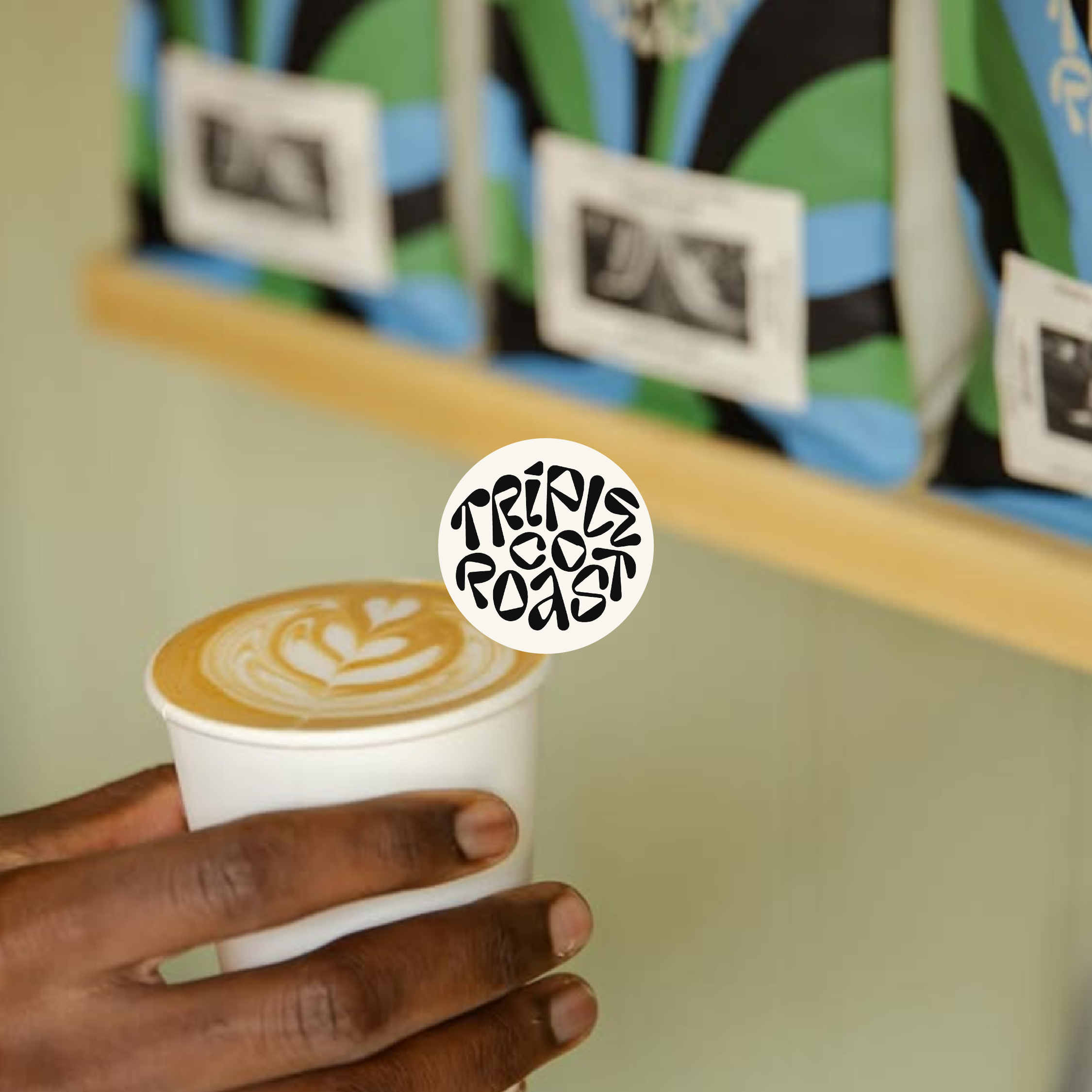 A person's hand holding a white paper cup with latte art, featuring a heart and leaf design, against a light green background.
