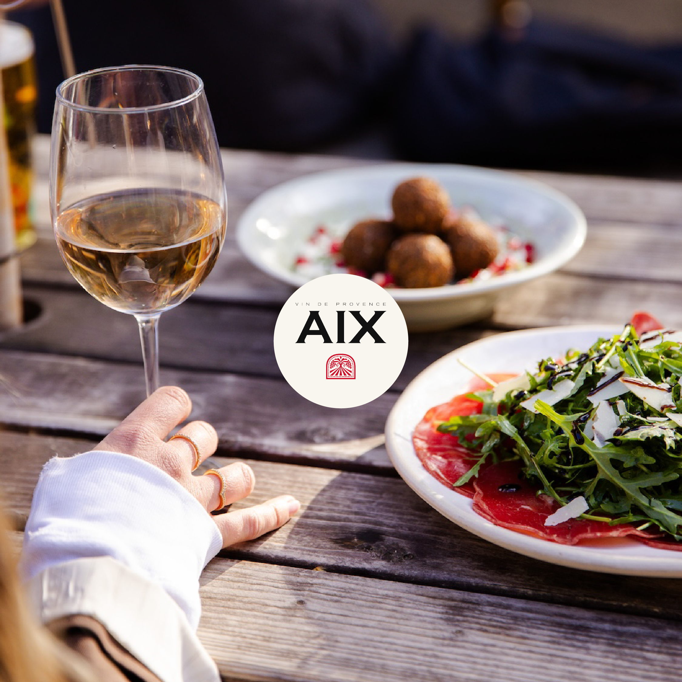 A person holding a glass of white wine with a plate of fried snacks and a salad on a wooden table outdoors.
