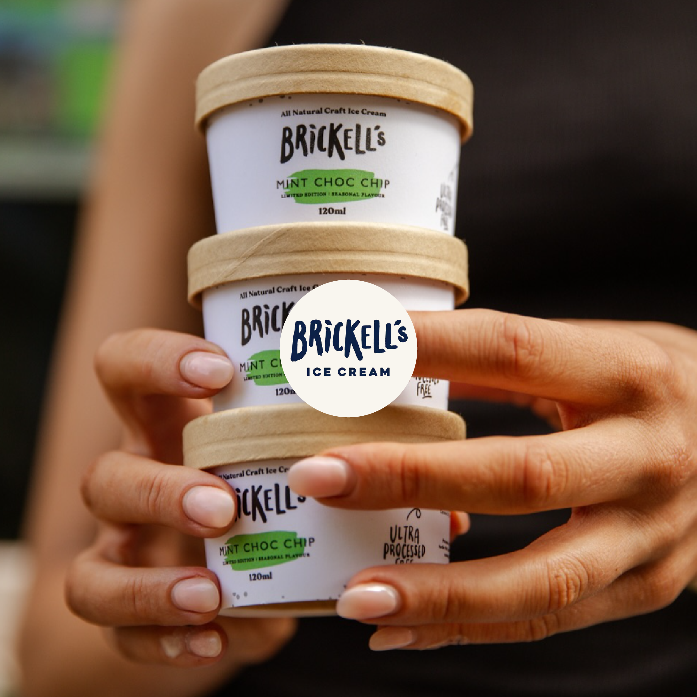 Close-up of a hand holding multiple Brickell's ice cream cones with a label that says Mint Choc Chip. The cones have a wooden look and the background is blurred.