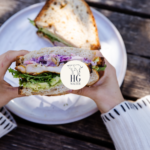 Person holding a sandwich filled with lettuce, purple coleslaw, and grilled chicken on a white bun, with a second sandwich on a white plate in the background, on a wooden table.
