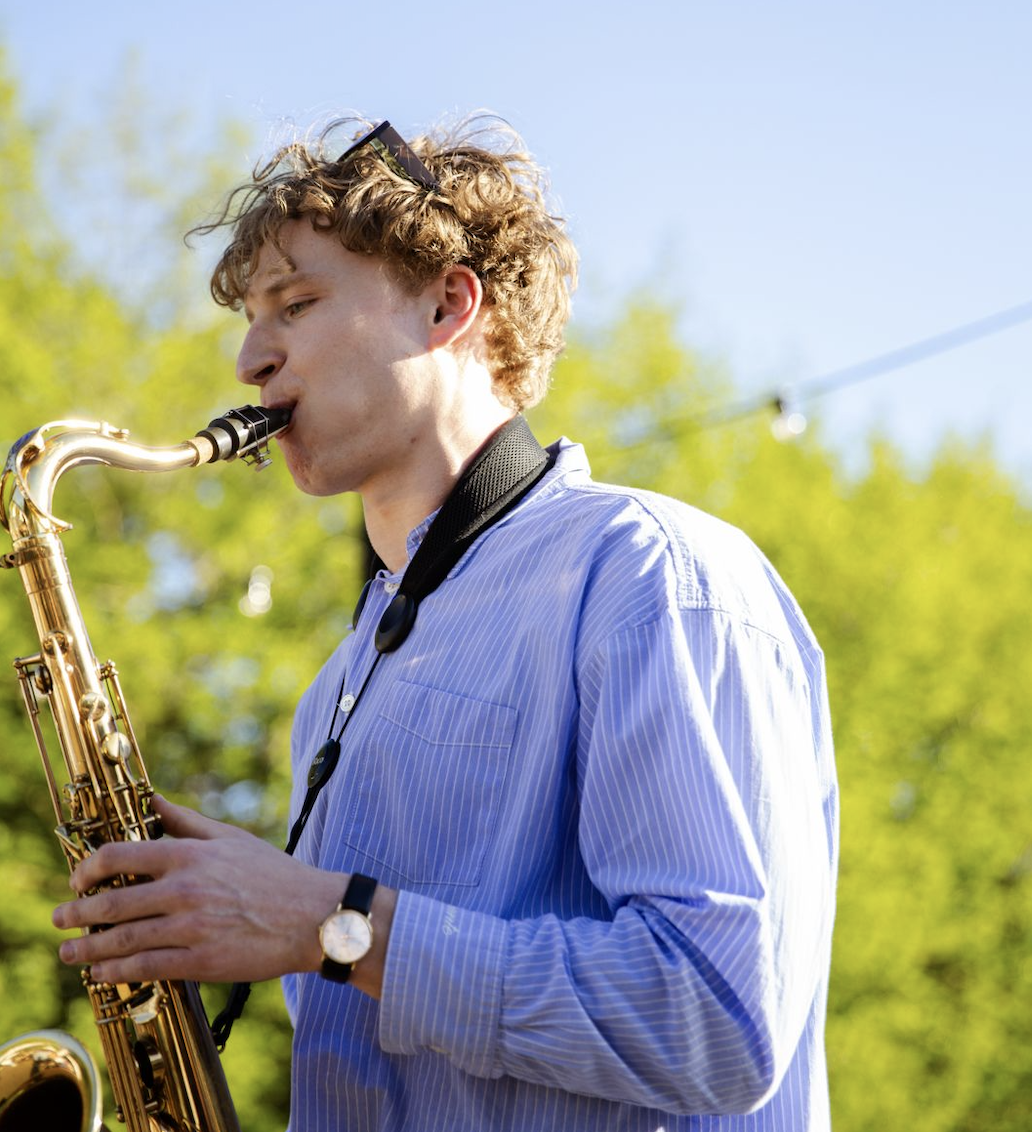 Young man with curly hair playing a saxophone outdoors, wearing a light blue dress shirt, a watch, and sunglasses on his head, with greenery and a clear sky in the background.