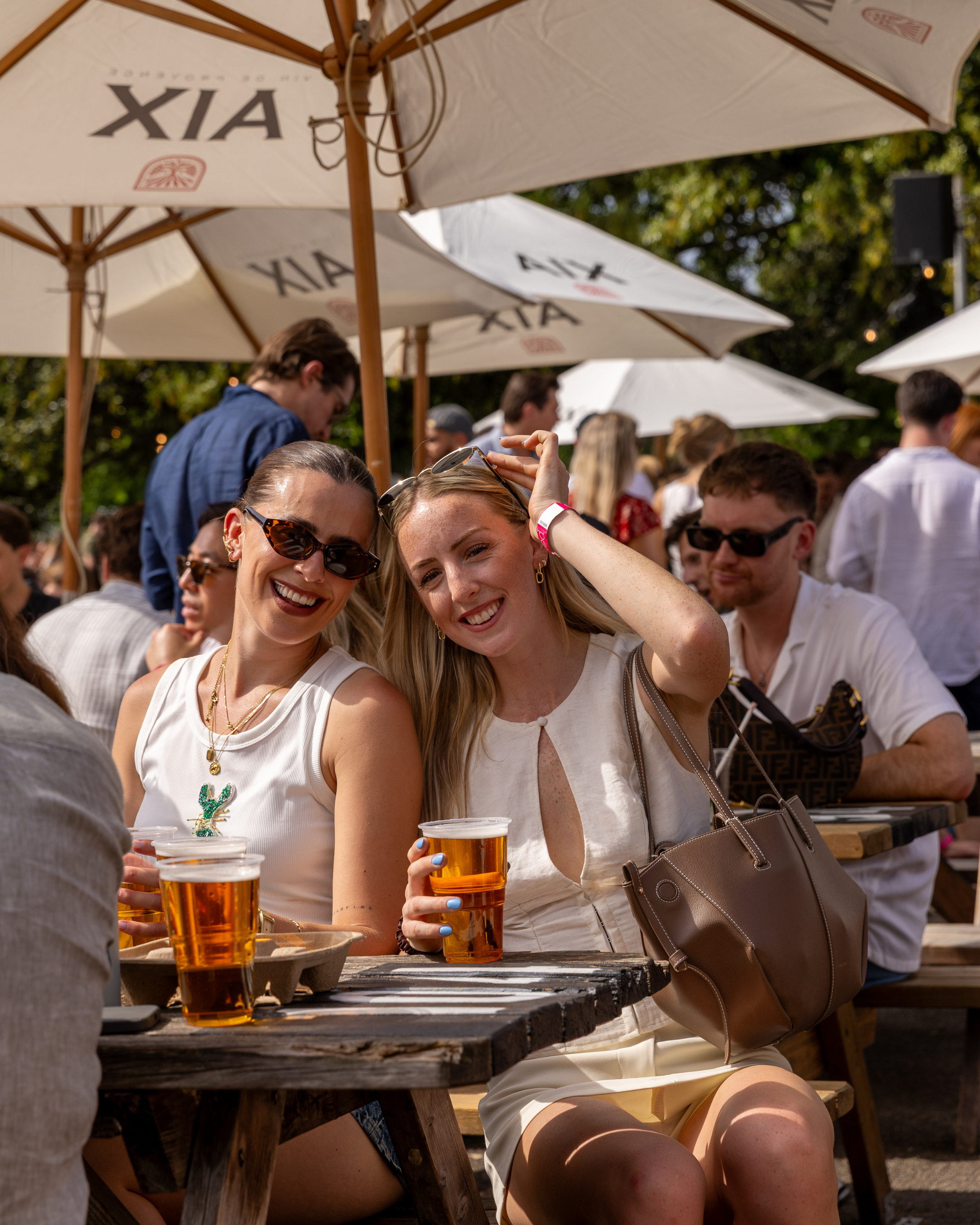 Close-up of smiling women holding pints of beer at an outdoor gathering under umbrellas with a crowd in the background.