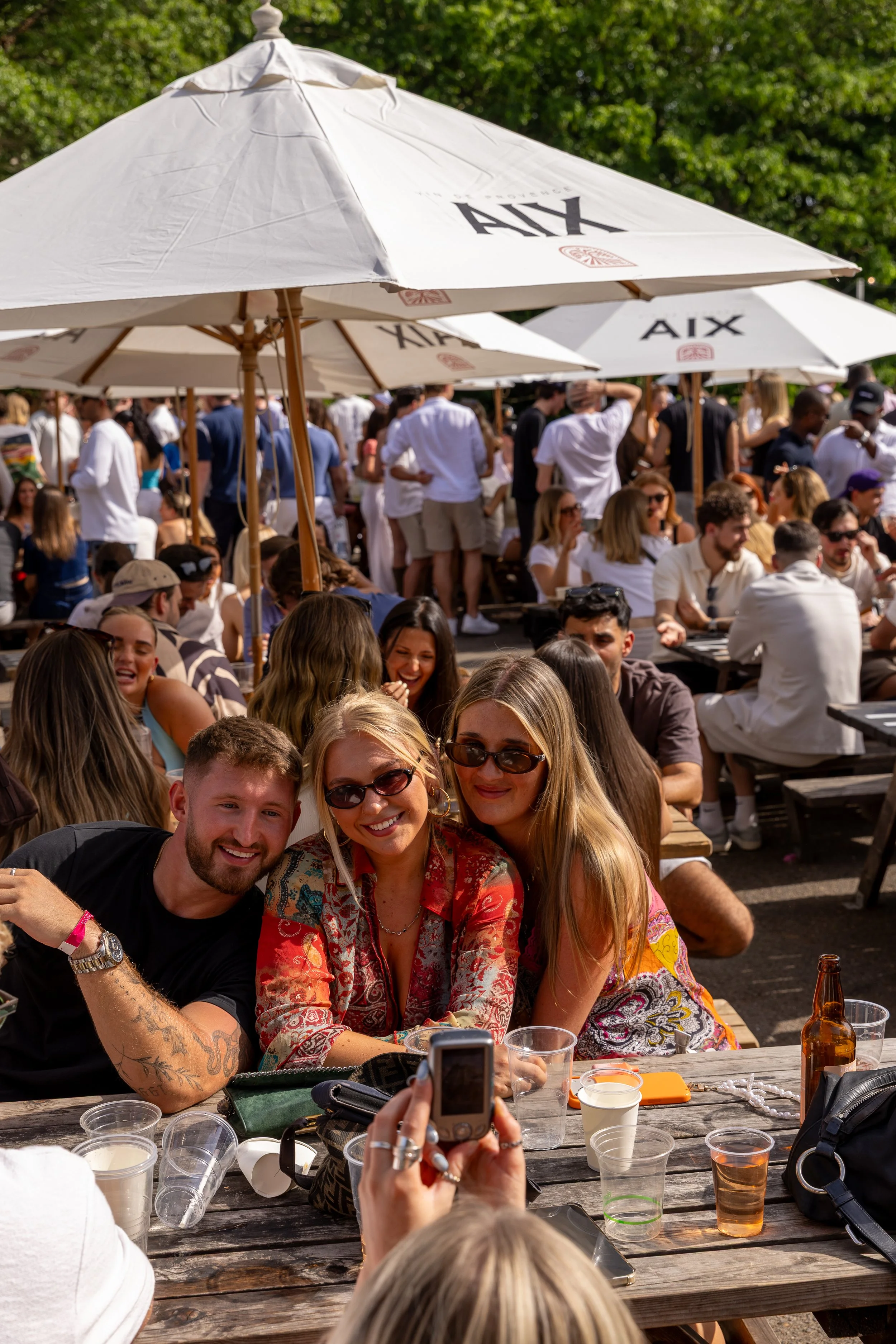 A group of people gathered outdoors under umbrellas, sitting at picnic tables, socializing, and taking photos on a sunny day.