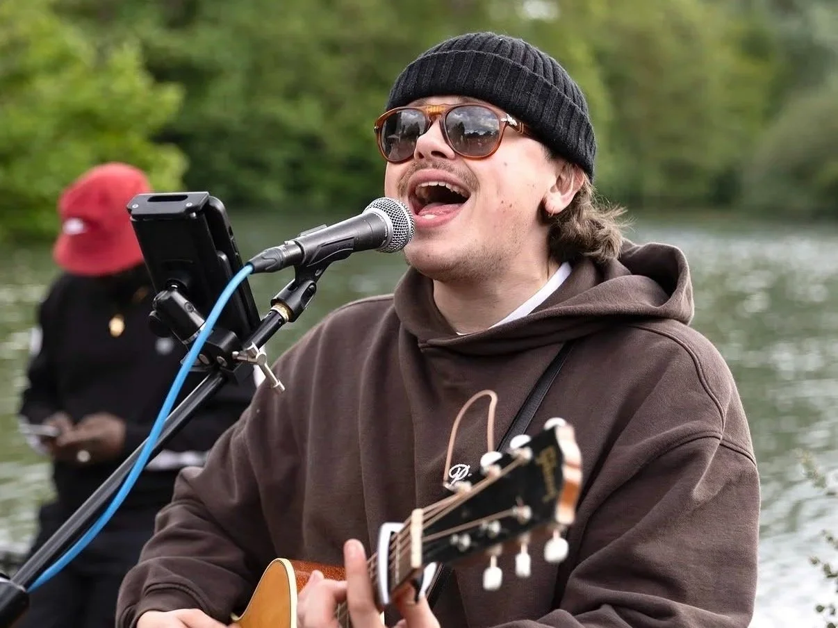 A man wearing sunglasses, a black beanie, and a brown hoodie playing an acoustic guitar and singing into a microphone outdoors near a body of water, with another person in the background.