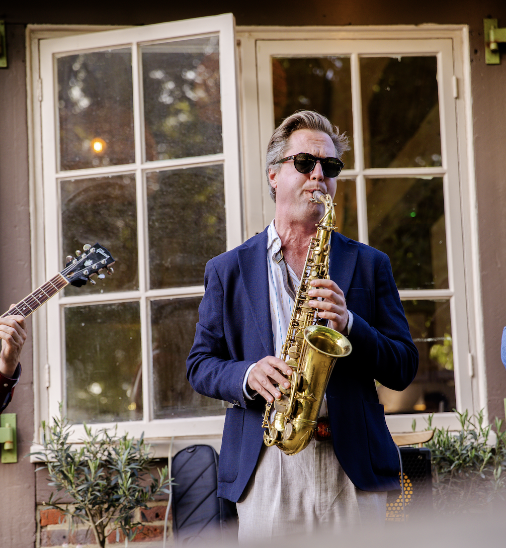 A man playing a saxophone outdoors with a window and plants in the background.