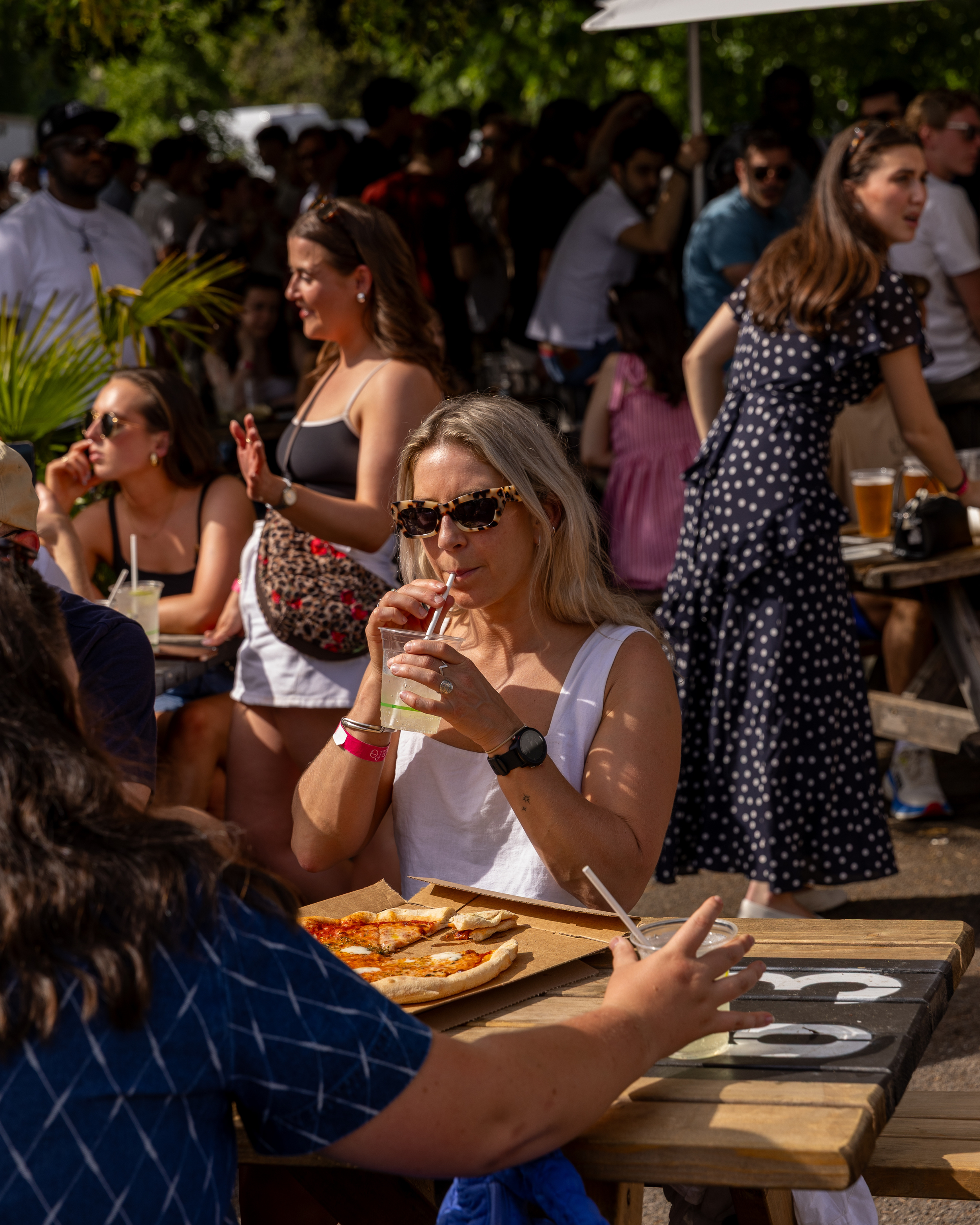 Group of people enjoying outdoor gathering with pizza and drinks, under trees with sunlight filtering through leaves.