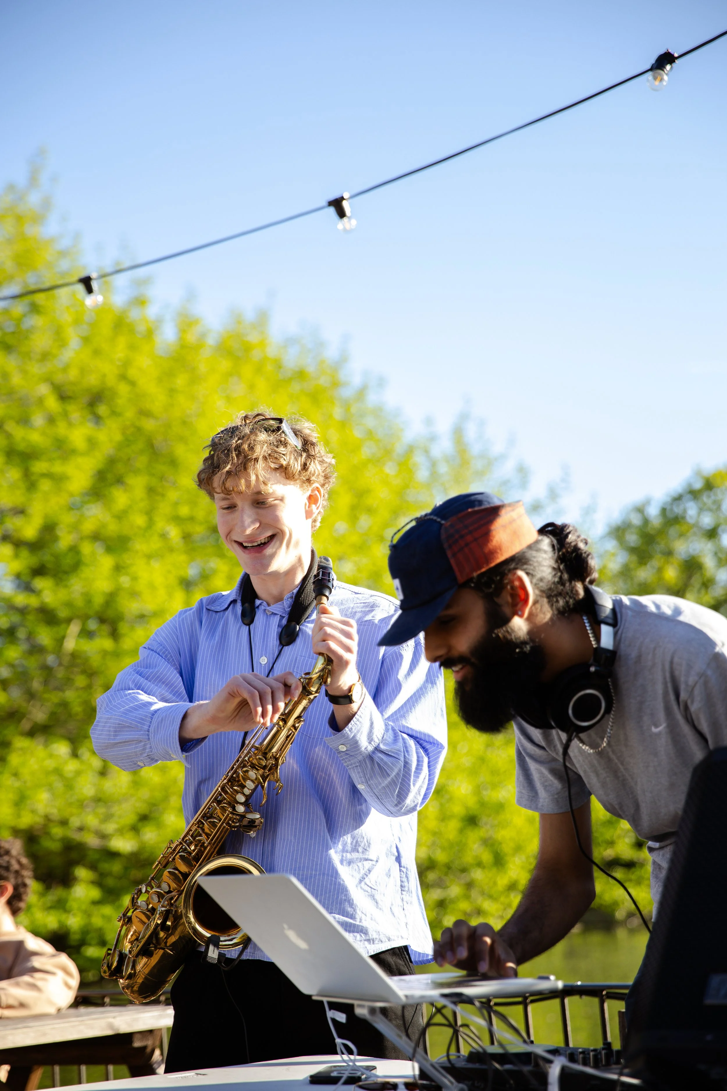 Young man playing saxophone outdoors, smiling, with two other men, one wearing headphones, in a park with trees and string lights overhead.