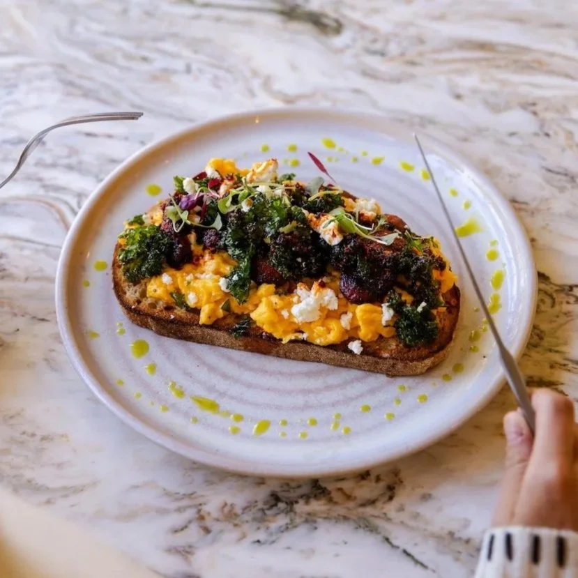 Close-up of a plate with a toasted bread topped with scrambled eggs, greens, and garnishes, on a marble table.