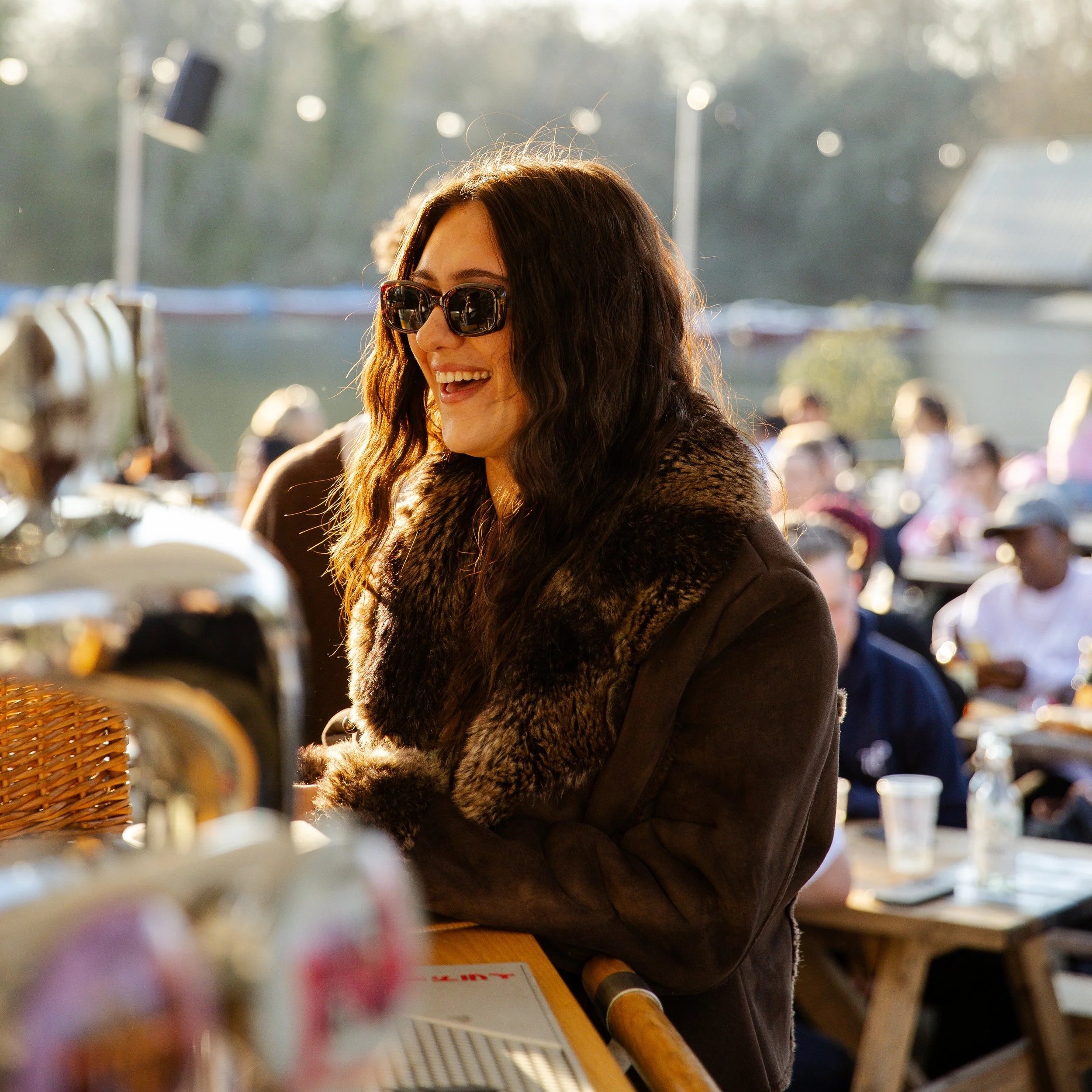 A woman with wavy brown hair wearing sunglasses and a fur-collared coat smiling in an outdoor setting with blurred people and trees in the background.