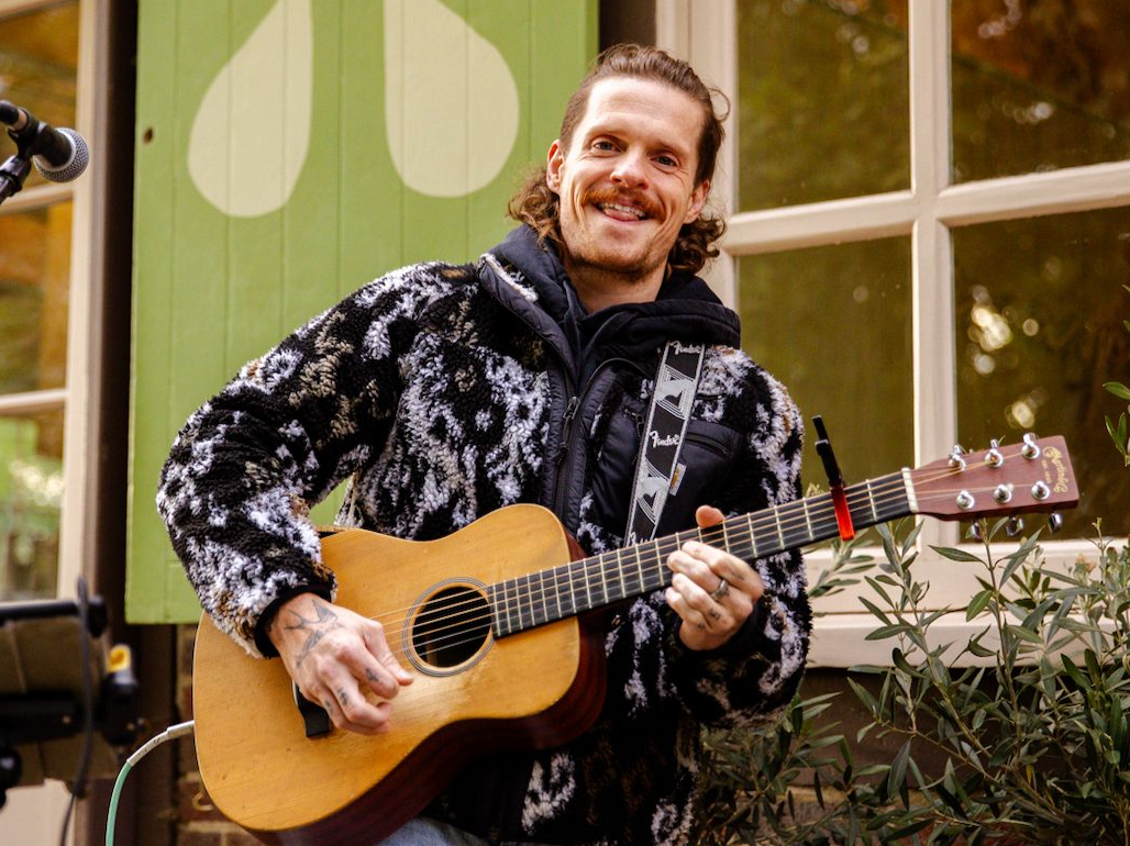 A man playing an acoustic guitar and smiling outdoors near a window and plants.
