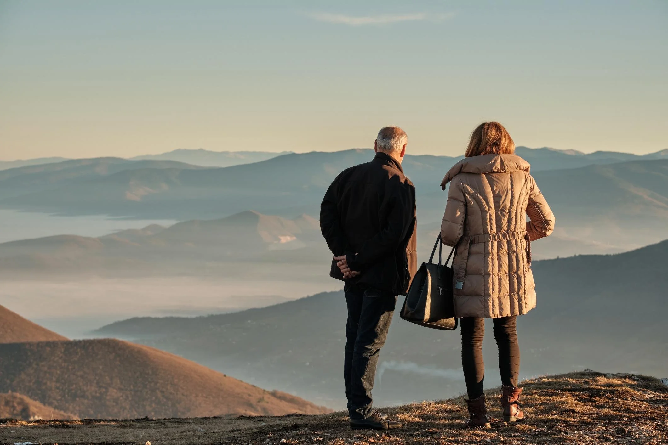 A man and a woman stand together overlooking a mountain landscape during sunset or sunrise, dressed warmly in jackets, with the man facing the mountains and the woman with her back to the camera.