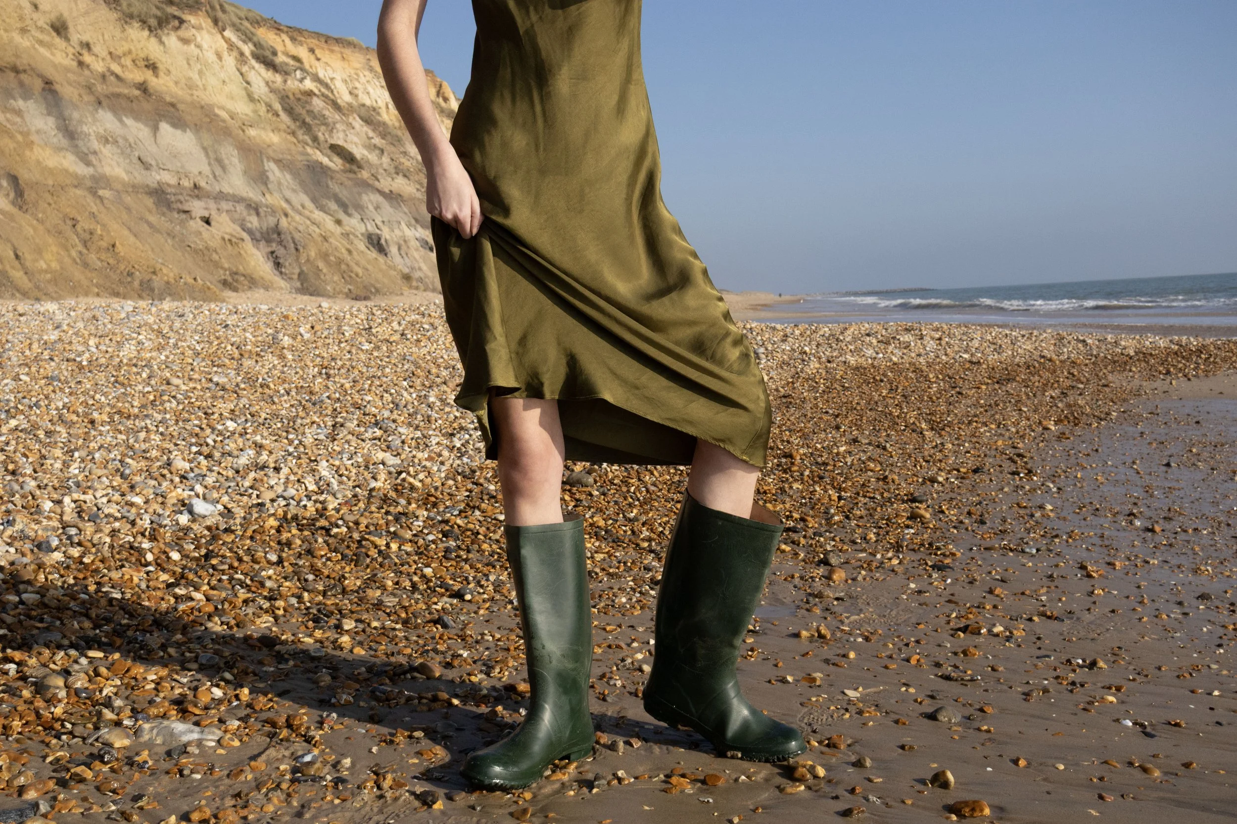 Person standing on a pebble beach near the ocean wearing a green dress and green rubber boots.