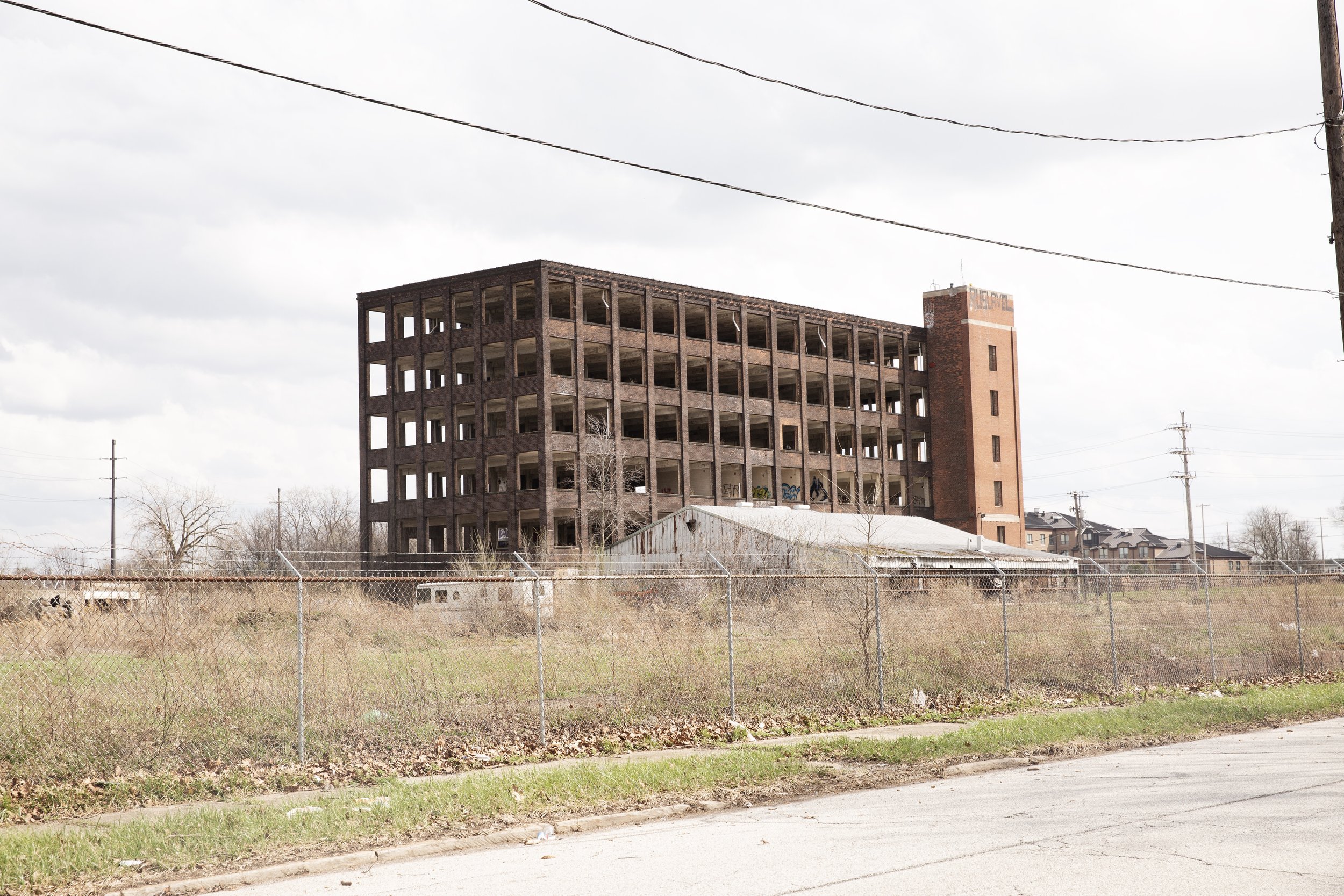An abandoned, multi-story brick building with open windows, surrounded by a chain-link fence and a grassy area, overcast sky.