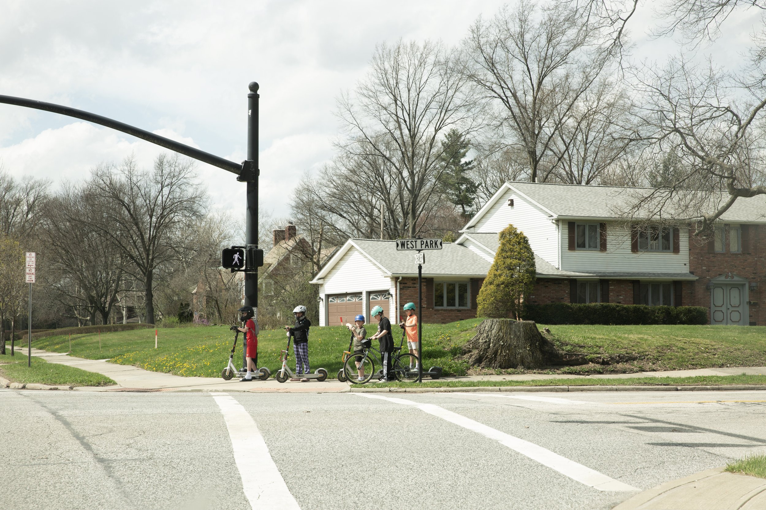 Children riding scooters and bikes at a crosswalk on a residential street in front of houses and leafless trees under a partly cloudy sky.