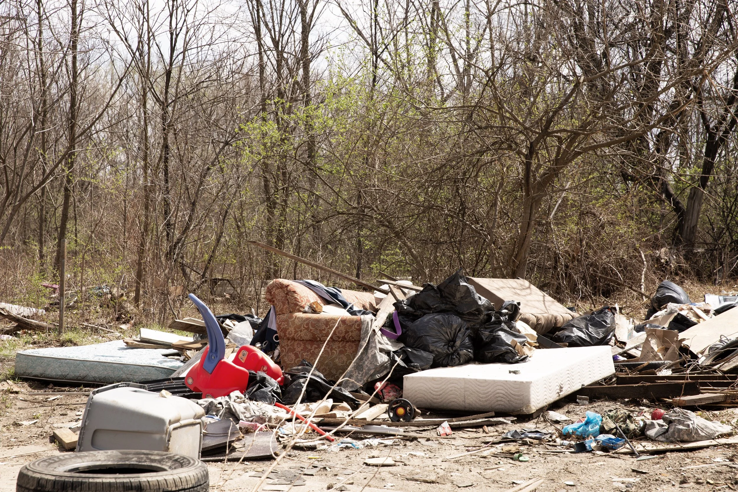 Trash and debris scattered on ground in front of leafless trees, including a mattress, black garbage bags, tires, a red child's ride-on toy, and household items.