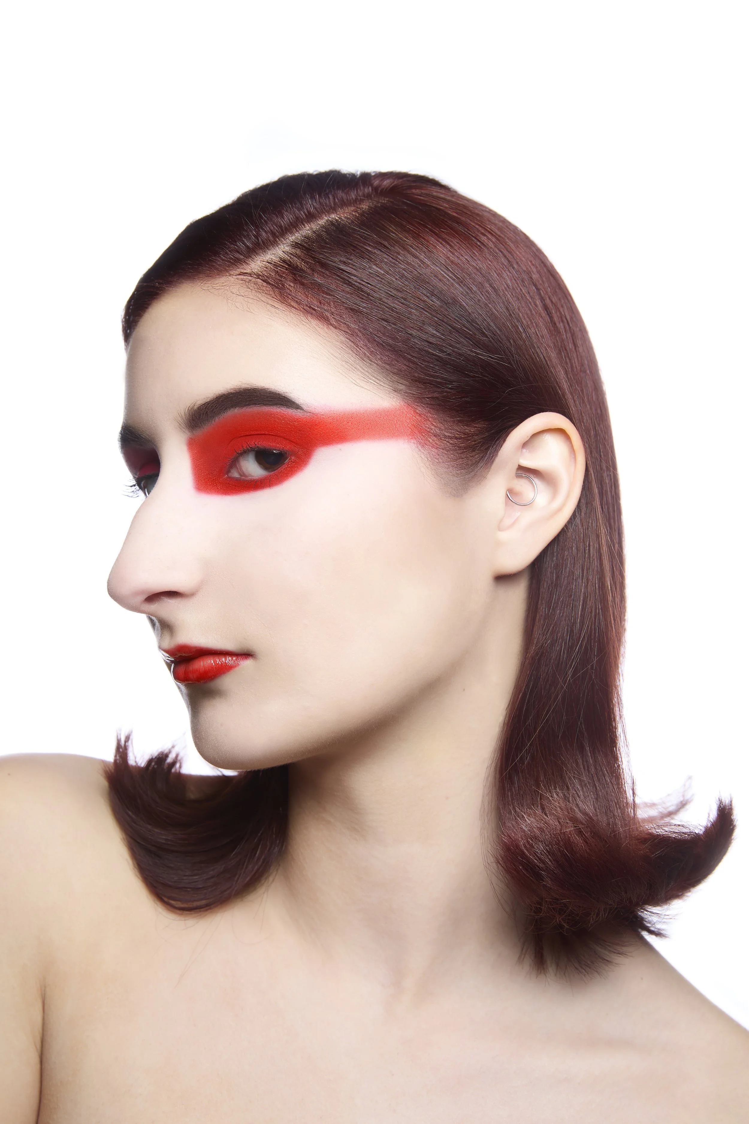 Close-up of a woman’s face with red hair styled in loose waves, wearing bold red eye makeup with thick eyeliner and matching lipstick, against a plain white background.