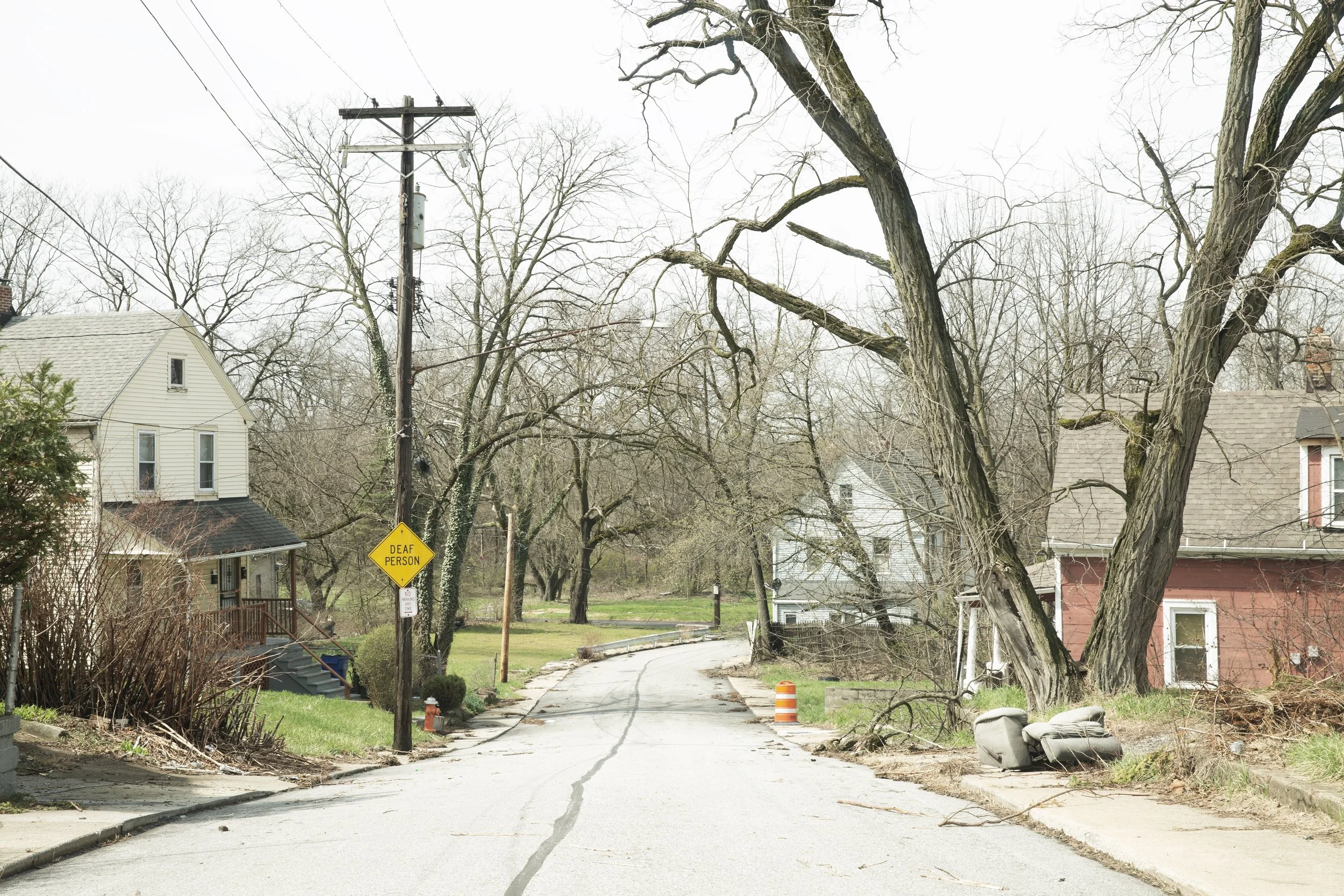 A suburban street with leafless trees, houses, utility poles, and a sign that reads "Deaf Person." The street appears damaged or under construction, with a traffic cone and debris visible.