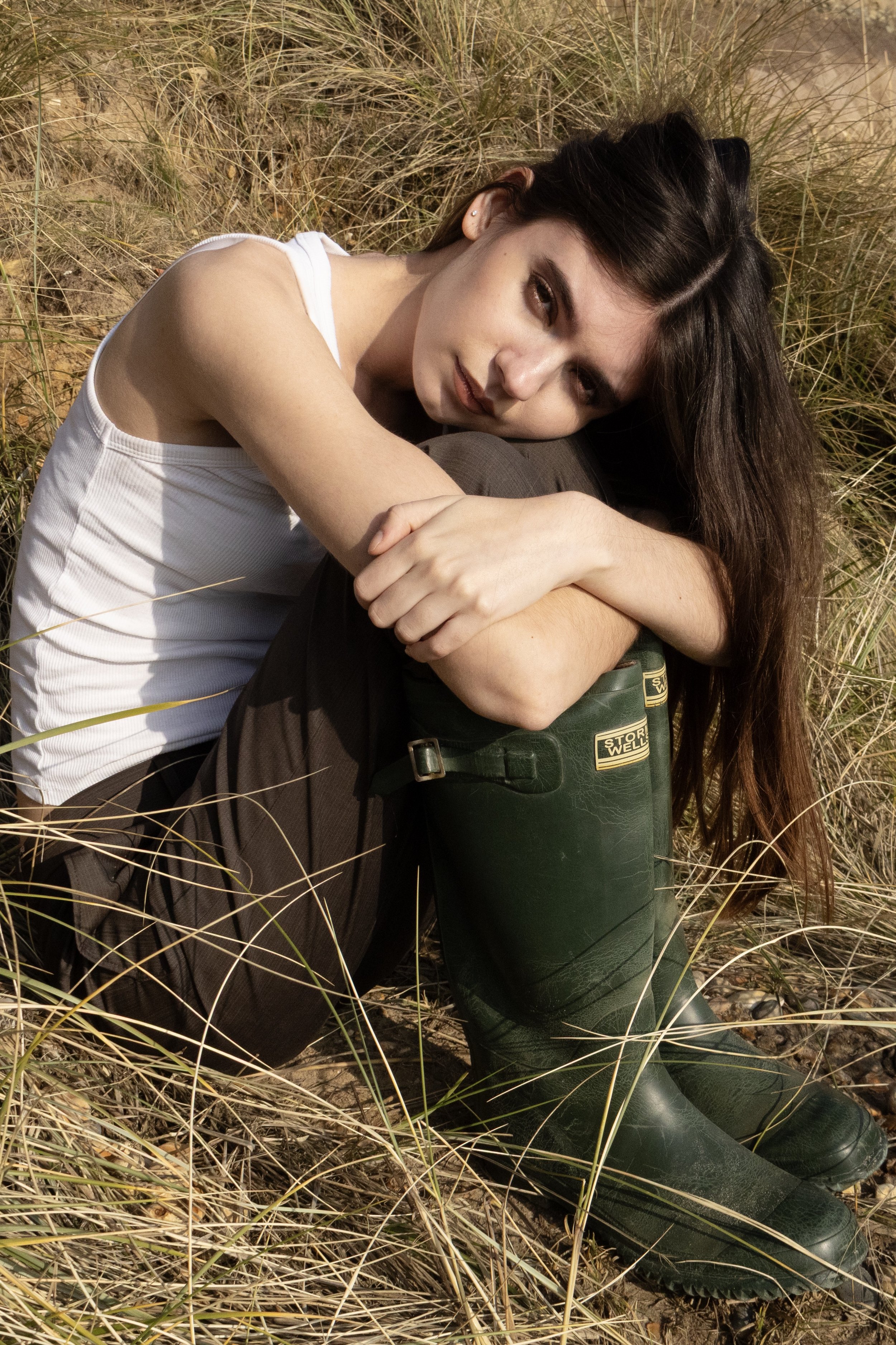 A woman with long brown hair lying on the grass in boots and a white shirt, outdoors during daytime.