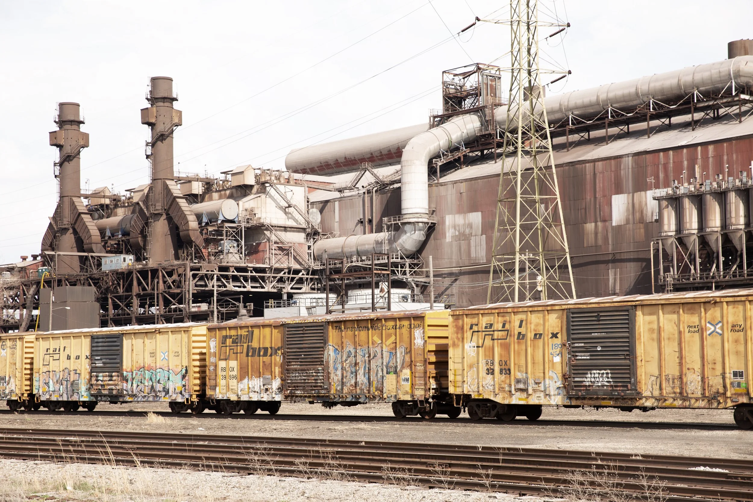 Industrial scene with rusted factory buildings, large pipes, and multiple freight trains with graffiti on yellow cars.