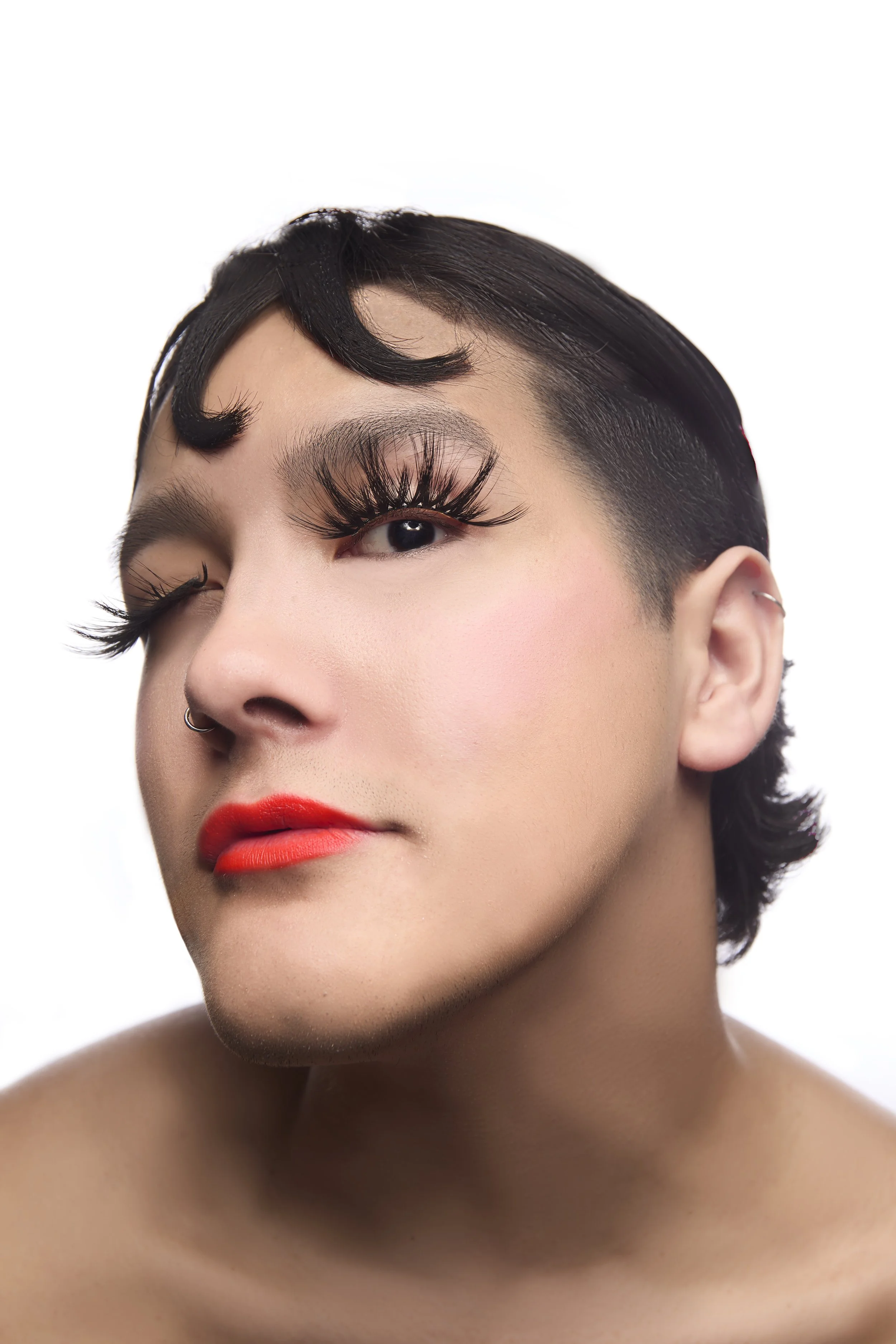 Close-up of a woman with short, dark hair styled with a curl, bold eyeliner and long eyelashes, red lipstick, and a nose ring, against a white background.