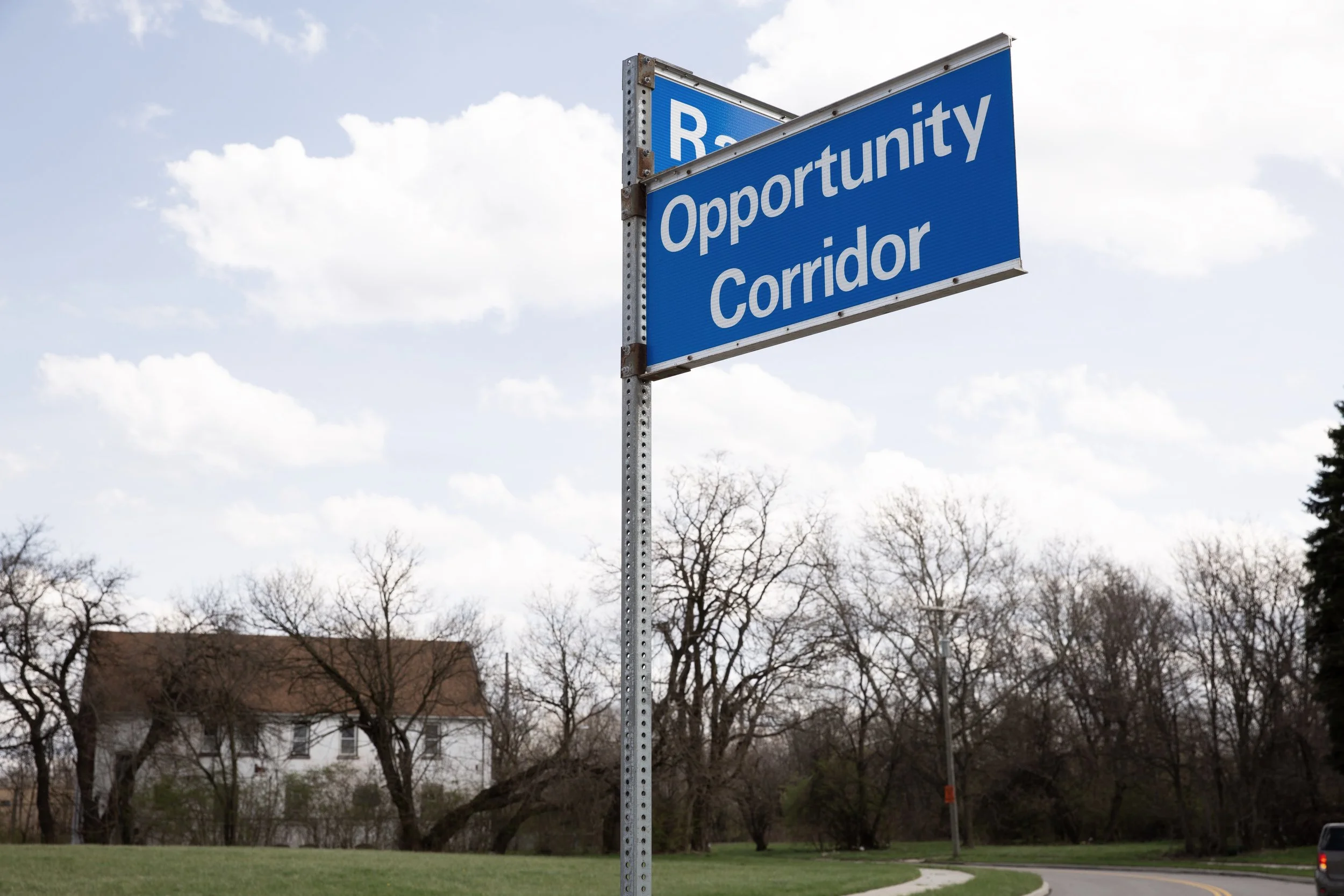 Blue street sign reading 'Race Opportunity Corridor' against cloudy sky, with trees and a building in the background.