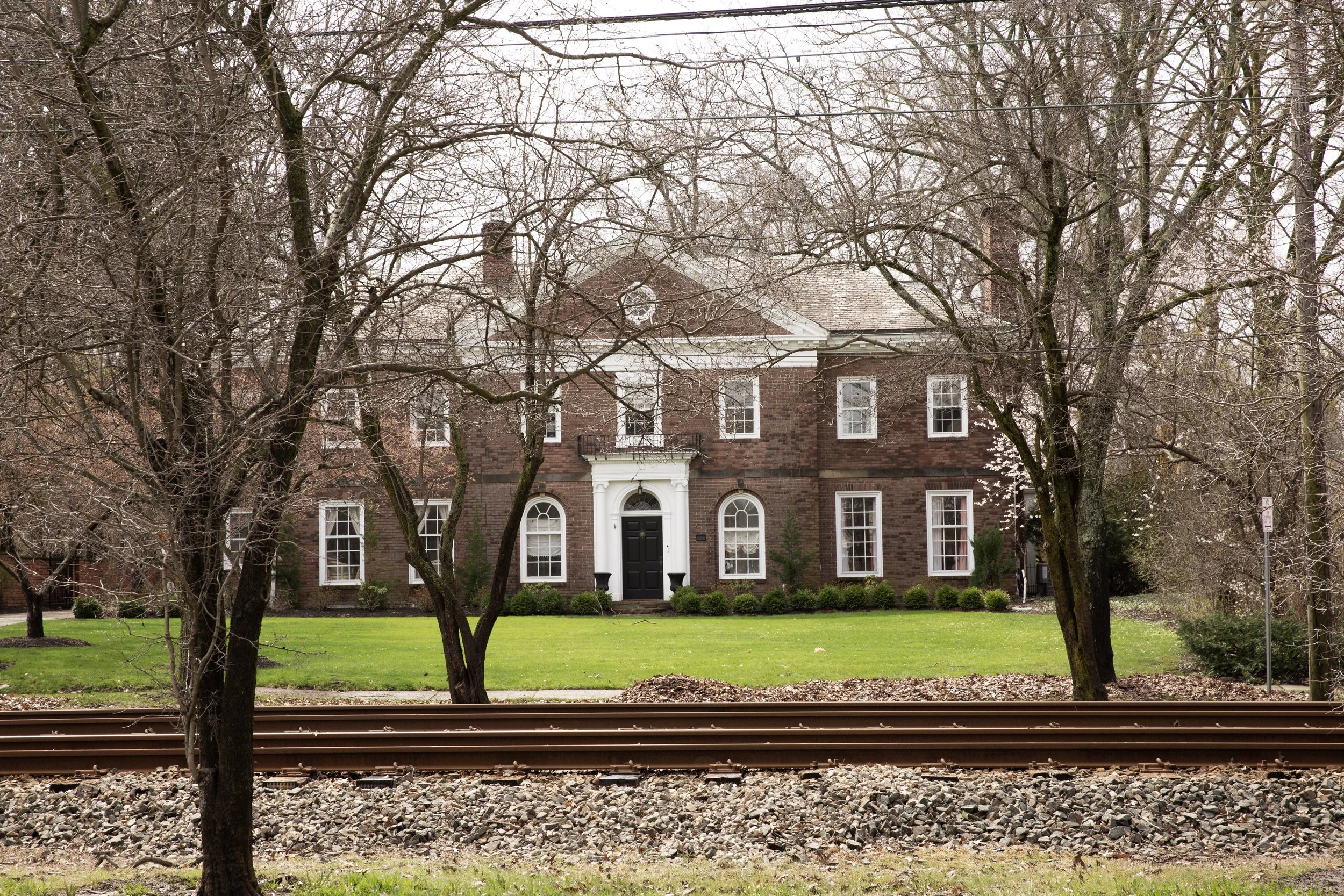 A brick house with white accents seen behind leafless trees and a grassy lawn, along with a railroad track in the foreground.