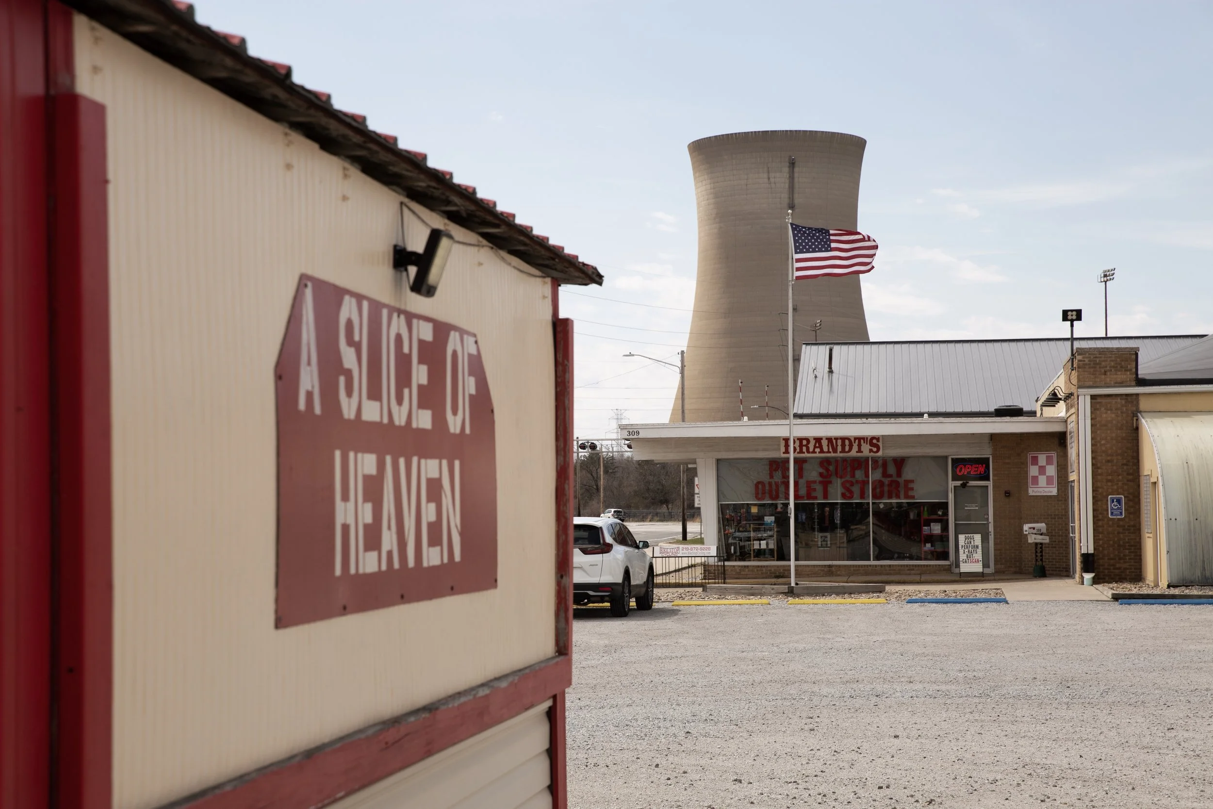 A red and white sign in the foreground reading 'A-1 SERVE HEAVEN,' with a pet supply store named 'BRANDT'S PET SUPPLY' visible in the background along with a parking lot, American flag, and a cooling tower in the distance.