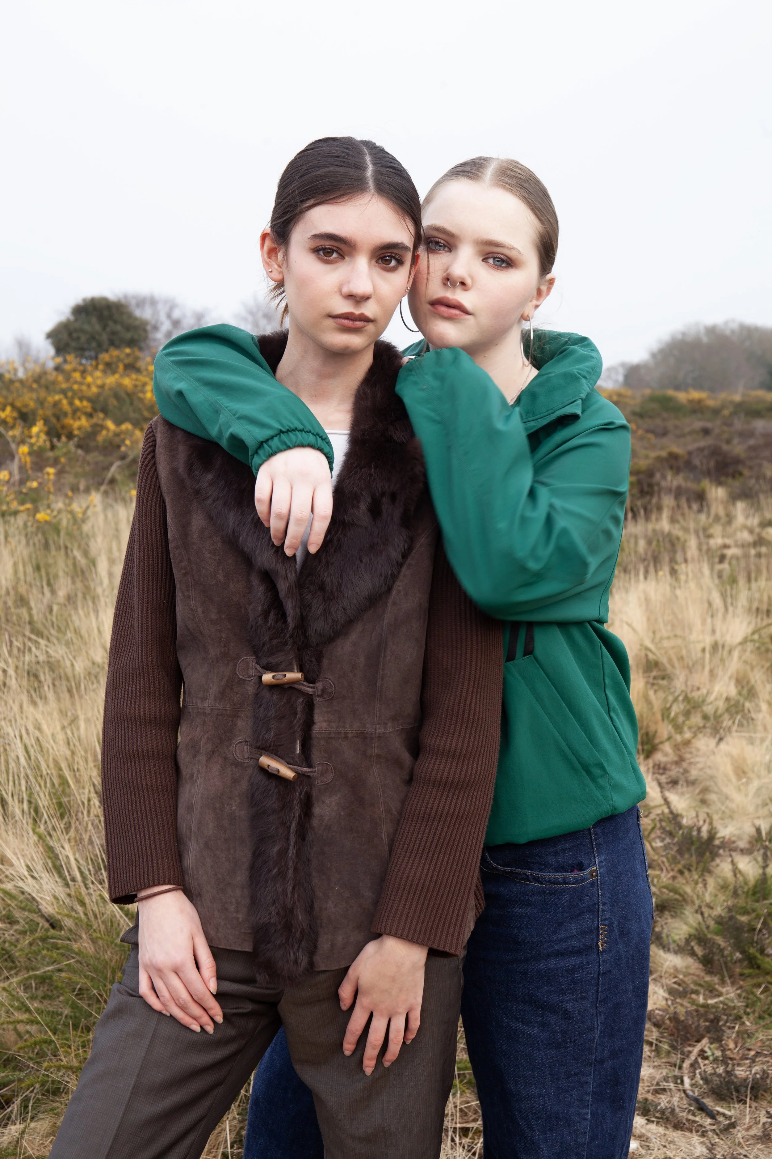 Two young women standing closely together outdoors, with one woman's arm around the other, in a field with grass and yellow flowers, overcast sky in the background.