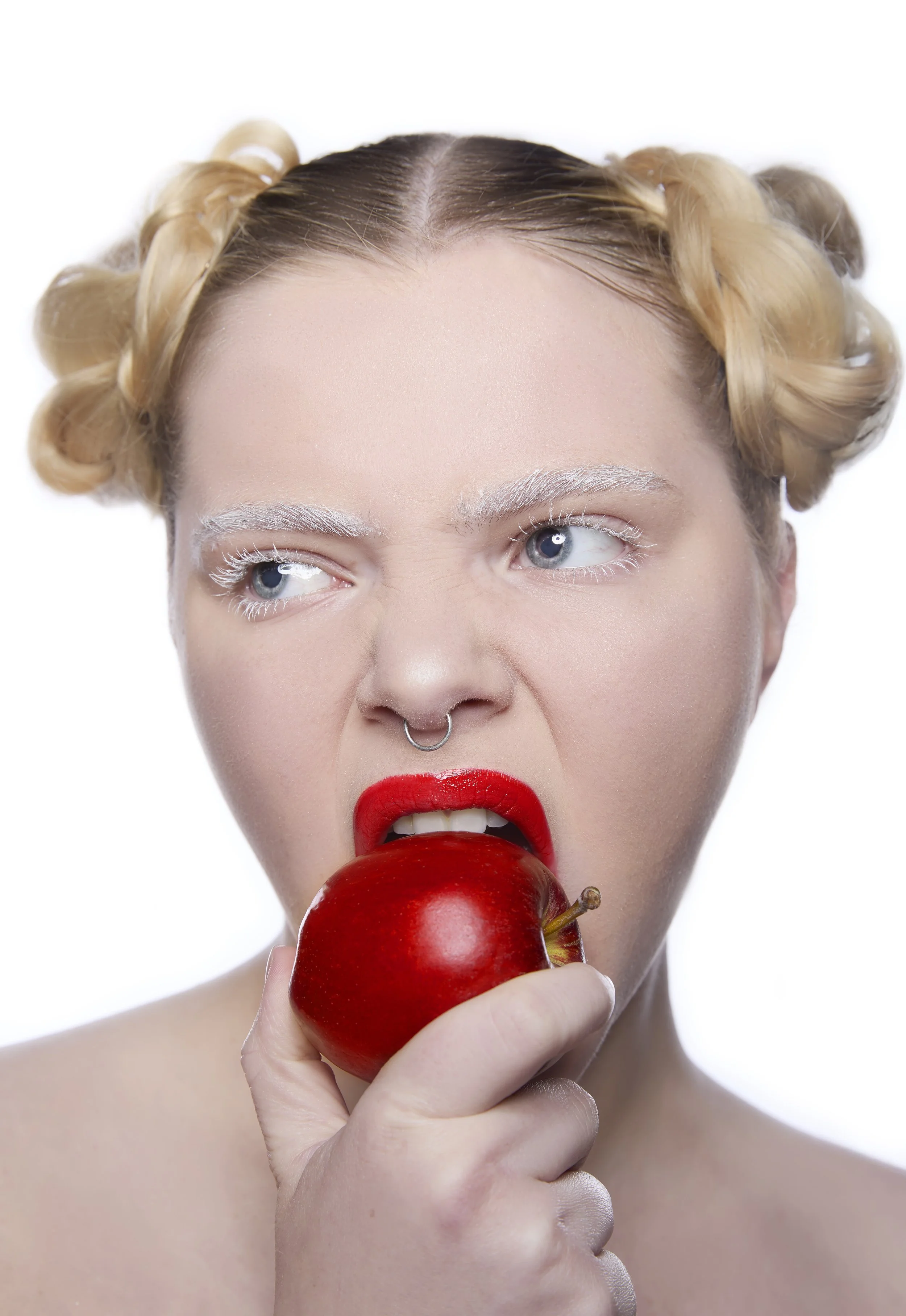 A woman with blonde hair styled in curls, wearing bright red lipstick and a nose ring, is holding a red apple near her open mouth.