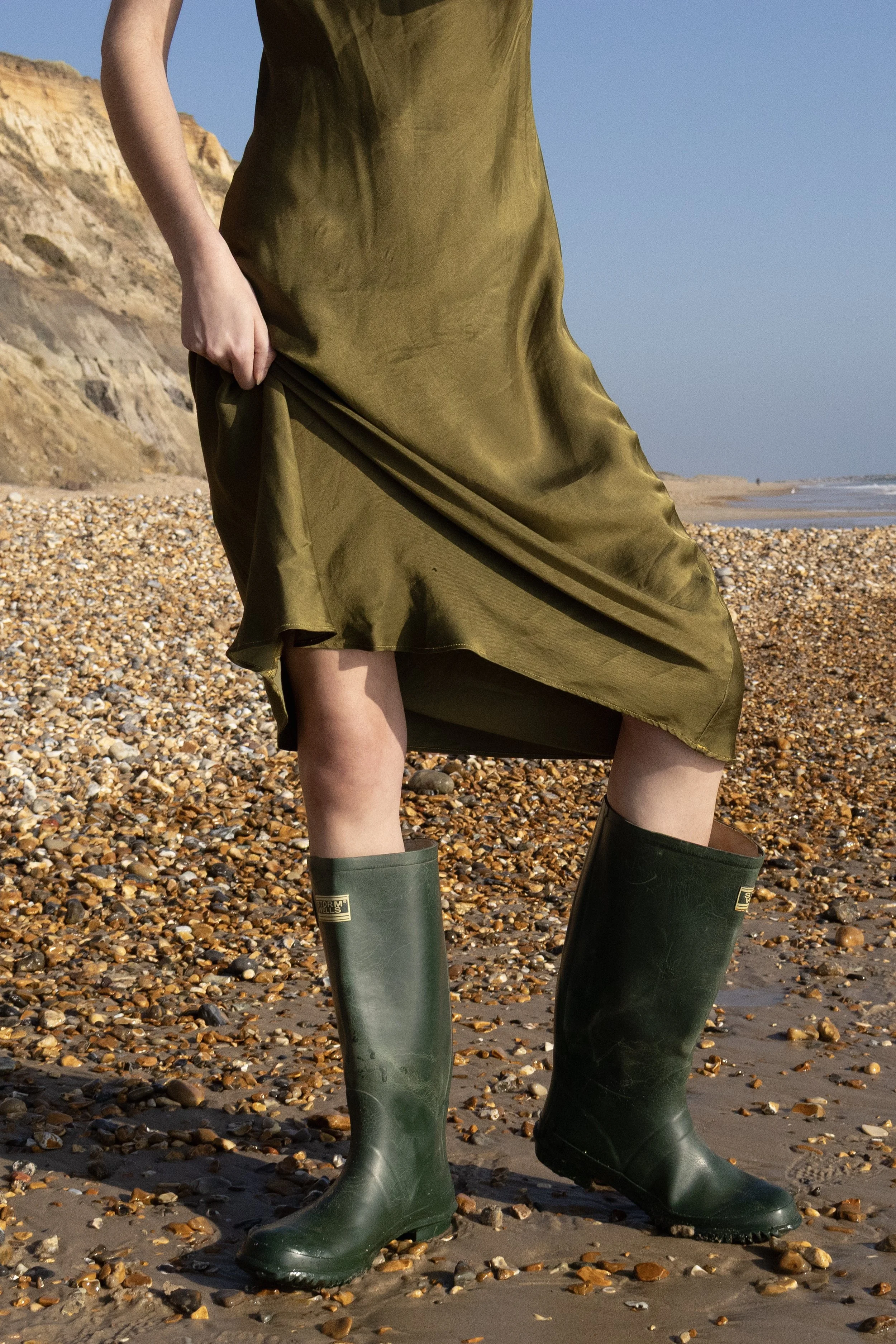 Person wearing a green dress and green rubber boots standing on a pebble-covered beach with cliffs in the background and a clear sky.