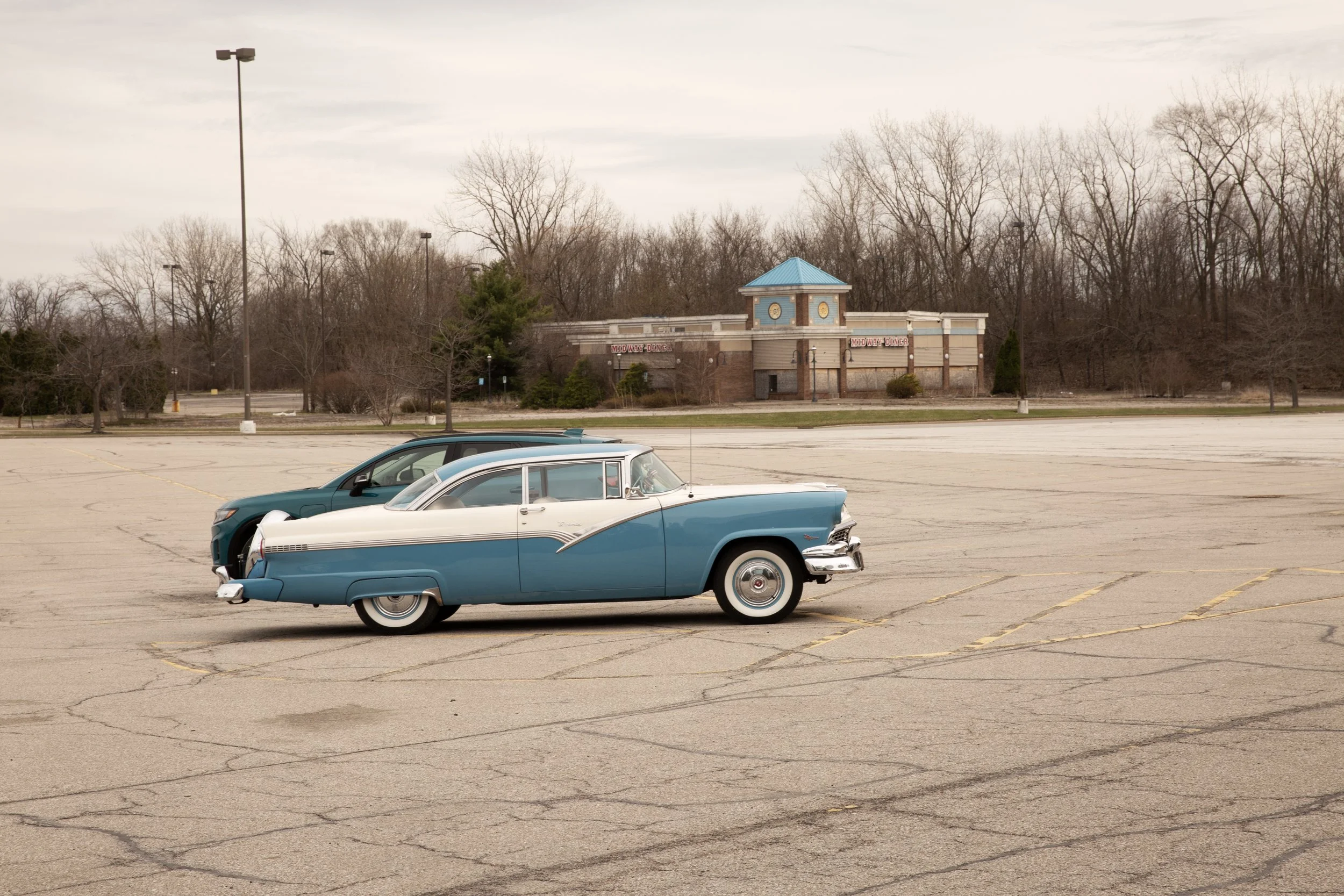 A vintage blue and white car parked in an empty parking lot with a Midway Diner in the background and leafless trees under a cloudy sky.