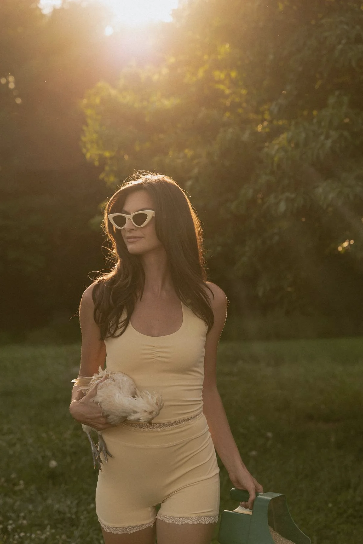 A woman with long, wavy brown hair and white cat-eye sunglasses walking outdoors in sunlight, holding a small white chicken, dressed in a yellow halter activewear top and matching shorts, with greenery and trees in the background.