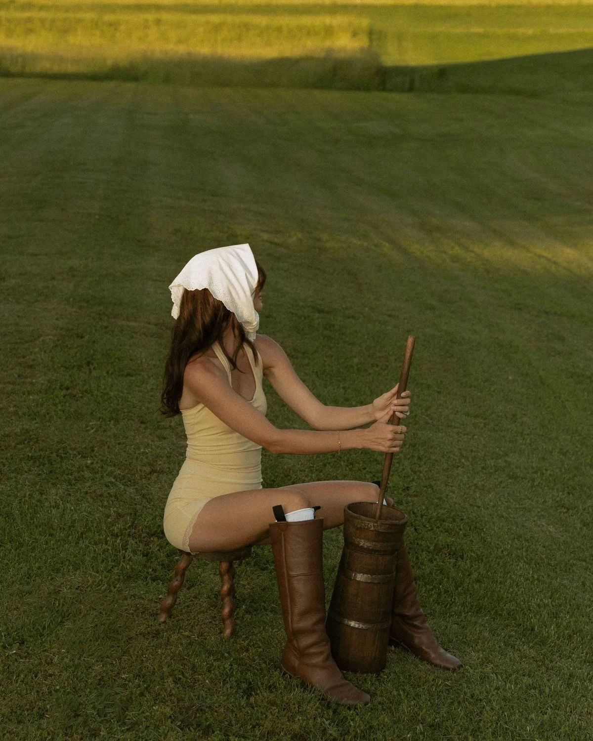 A woman sitting on a small wooden stool in a grassy field, stirring a large wooden barrel with a long stick, wearing a yellow halter top, matching shorts, and a white cloth on her head, with tall brown boots next to her.