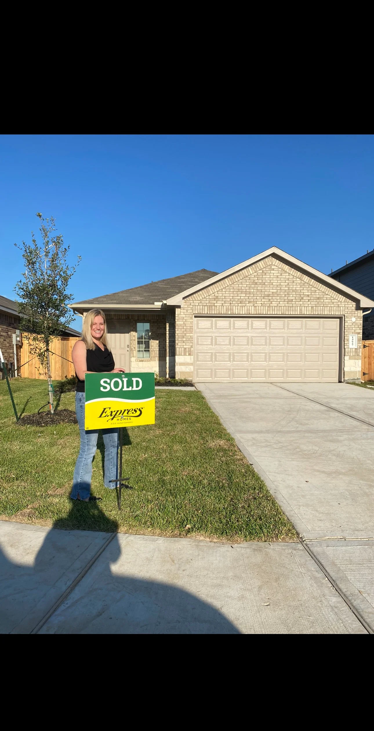 A woman standing on a lawn holding a green and yellow sign that reads 'SOLD' in front of a new brick house with a two-car garage under a clear blue sky.