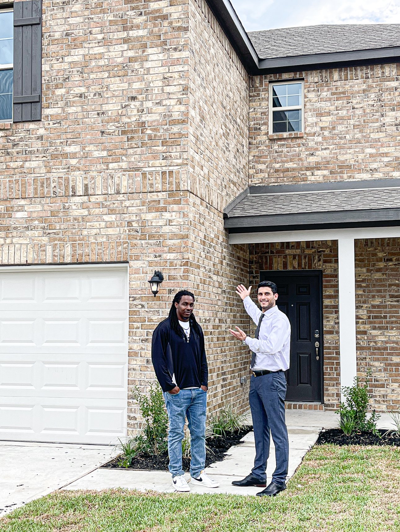 Two men standing in front of a brick house, one in a white shirt and tie gesturing towards the house, the other in a black jacket and jeans, looking at the camera.