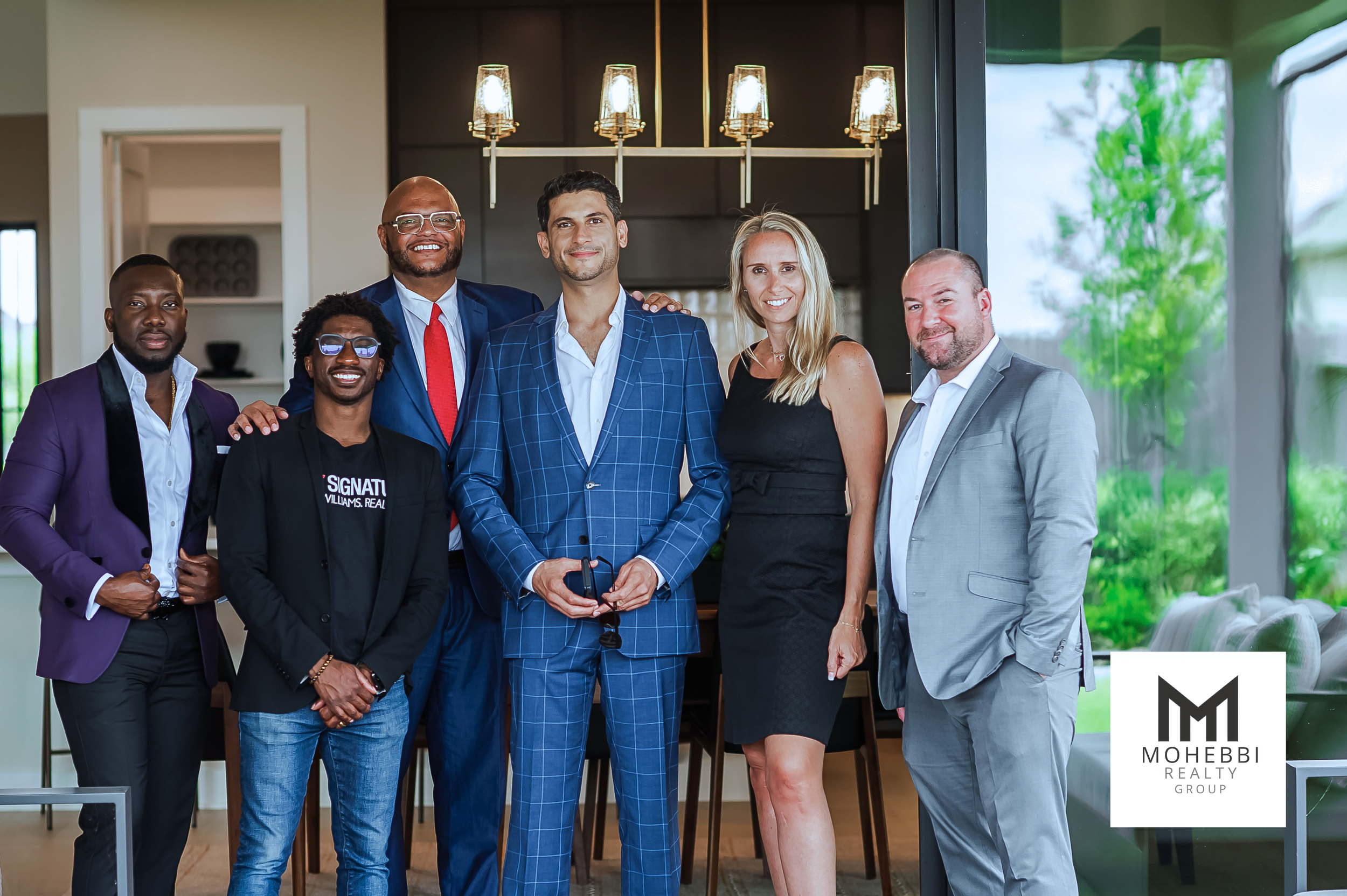 Group of six diverse professionals standing together in modern home interior, smiling at the camera, with MOHEBBI REALTY GROUP logo in the bottom right corner.