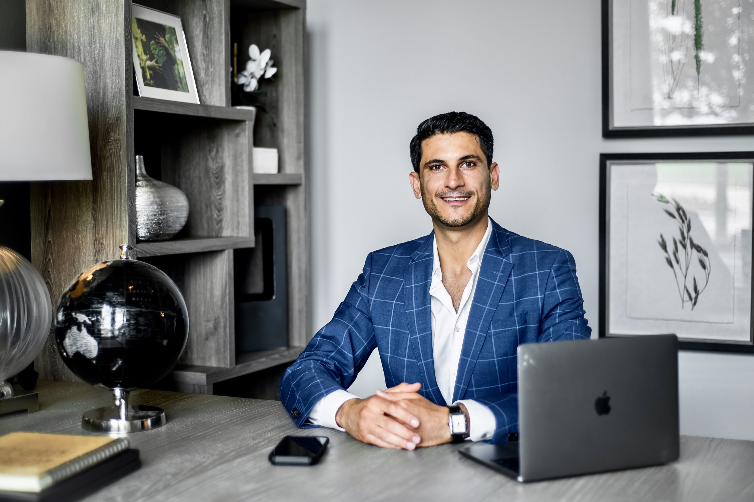 A man in a blue checkered suit sits at a desk in an office, smiling at the camera. The desk has a laptop, smartphone, and notebook. Behind him are framed artworks and a wooden bookshelf with decorative items and a white floral figurine.