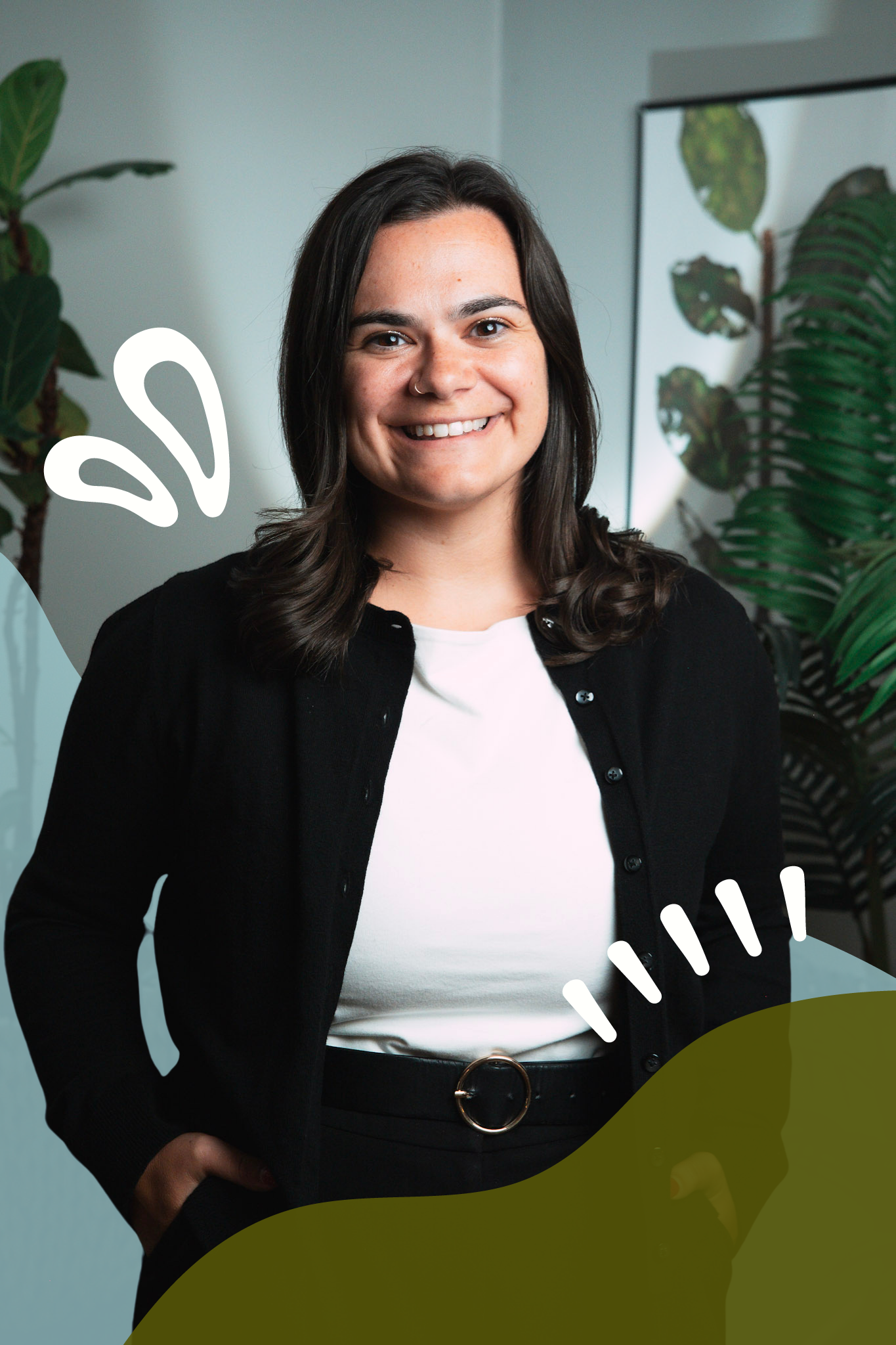 A woman with dark brown hair smiling at the camera, wearing a black cardigan, white shirt, and black belt, standing indoors with green plants and a mirror in the background.
