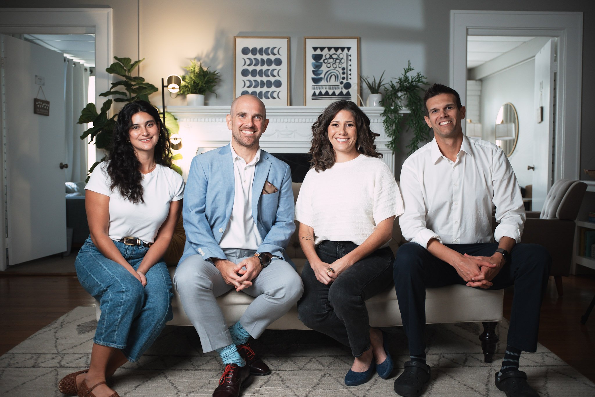 Five people sitting on a couch smiling at the camera in a living room with plants and framed art in the background.