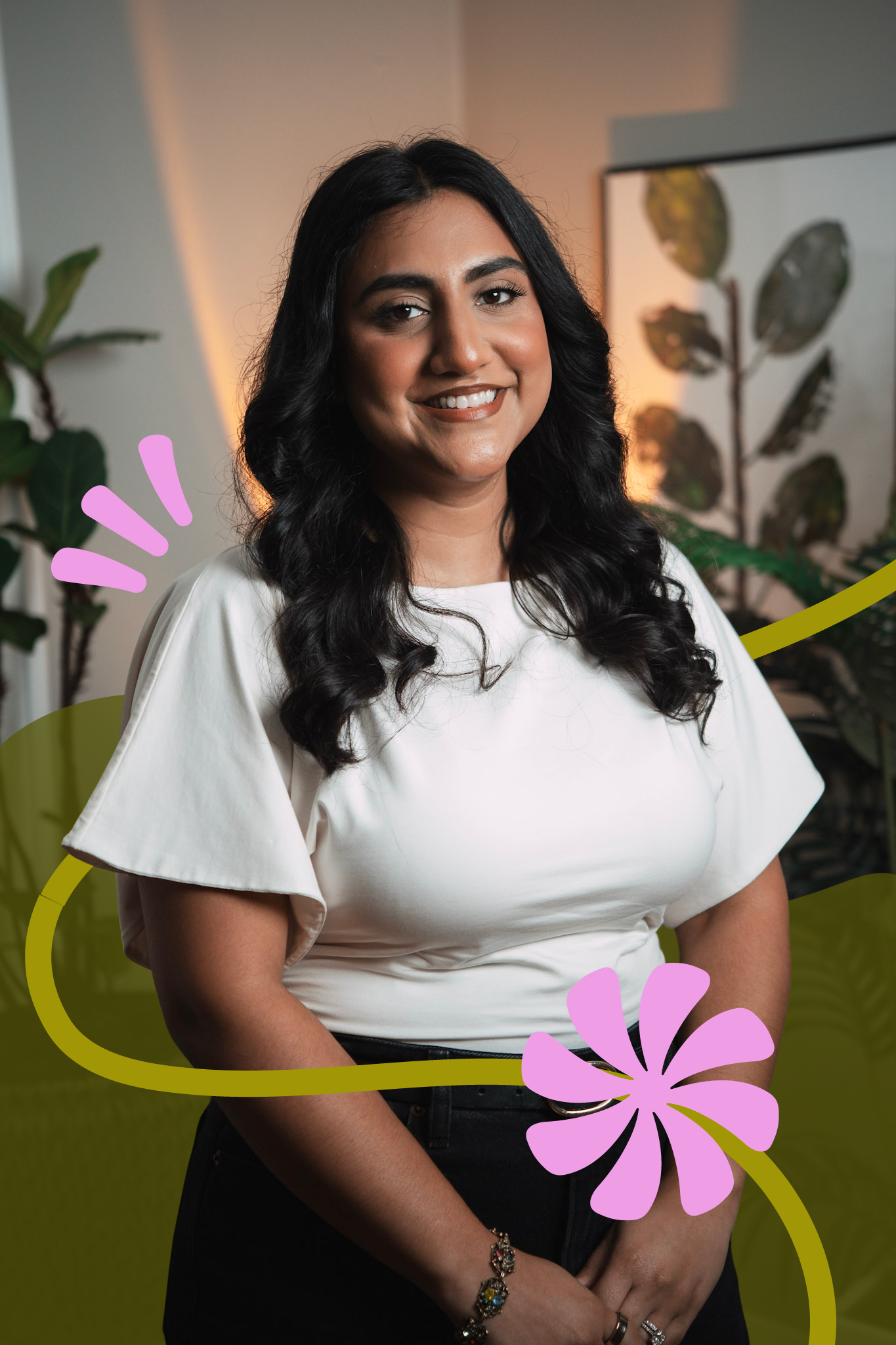 A woman with long, dark curly hair, smiling, wearing a white top with ruffled sleeves, standing in front of indoor plants with blurred background.