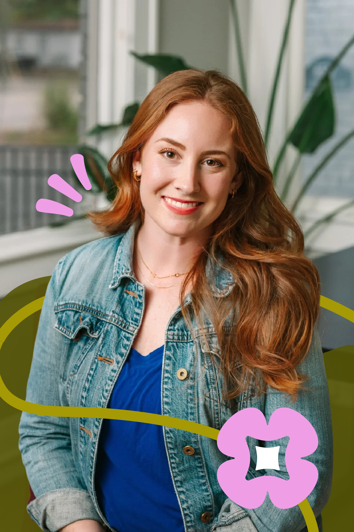 Young woman with long red hair smiling inside a bright room with large windows and green plants, wearing a denim jacket and a blue top.