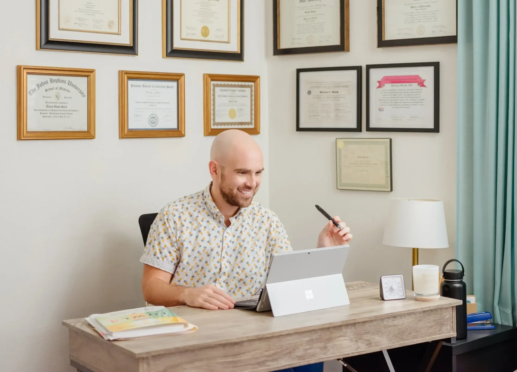 A man with a beard and a bald head, wearing a white shirt with colorful dots, is sitting at a desk in an office. He is smiling and holding a pen. The desk has a laptop, a tablet, books, a clock, and a coffee mug. The wall behind him is decorated with multiple framed certificates and diplomas.