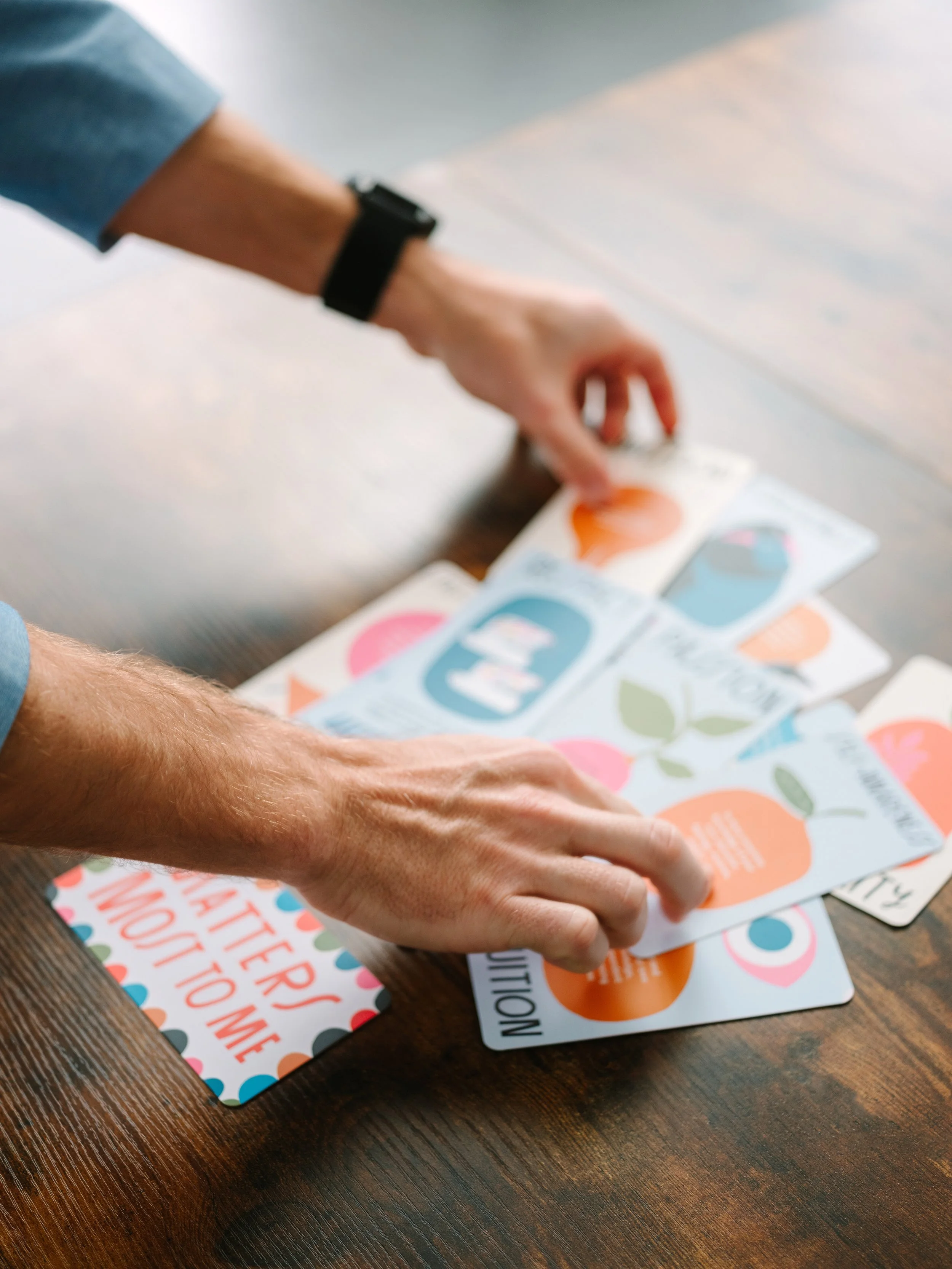 Person with light skin and a black wristwatch sorting through colorful cards on a wooden table.
