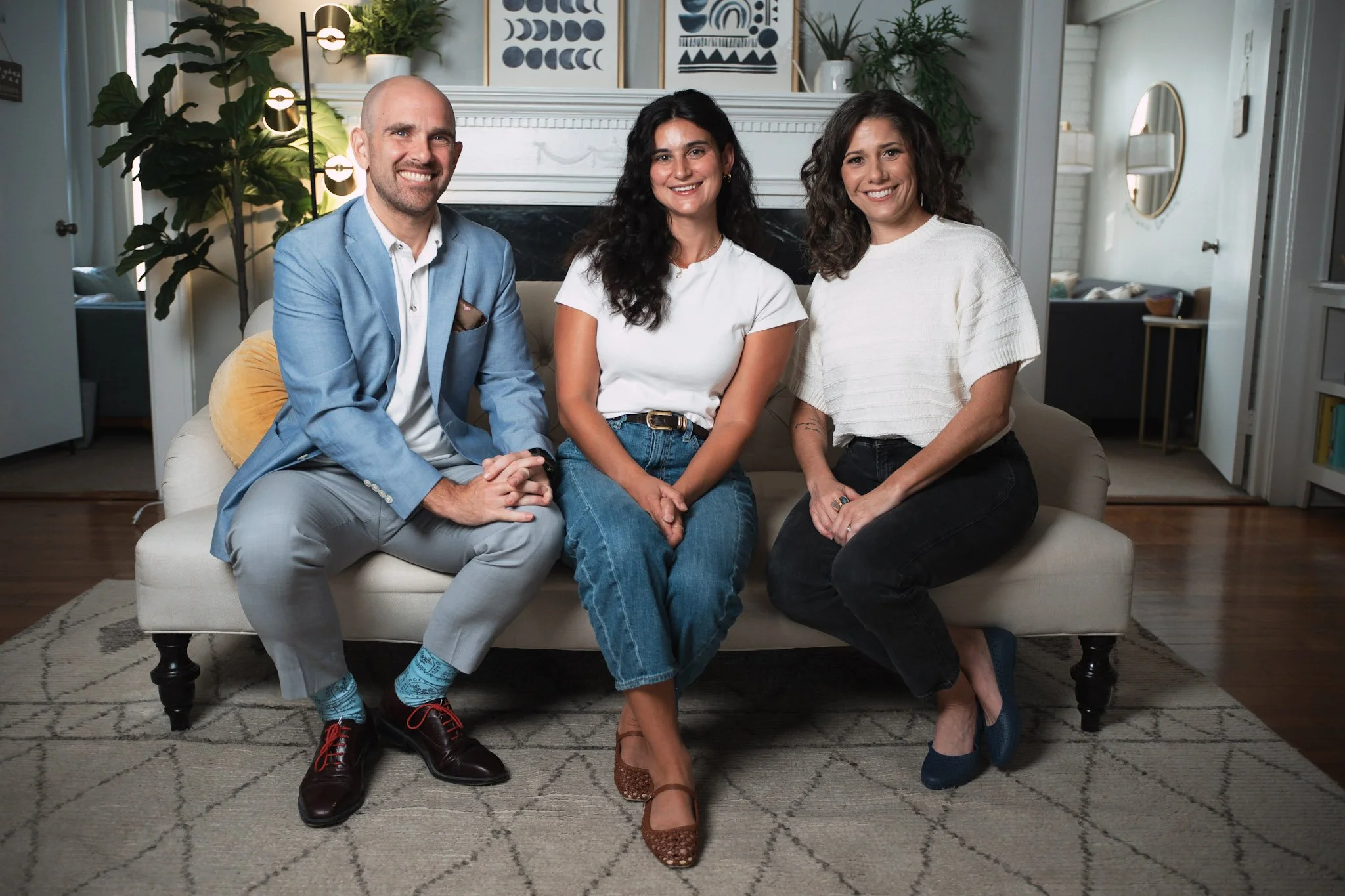 Three people sitting on a white sofa in a cozy living room, smiling at the camera. The man on the left is wearing a light blue blazer, white shirt, and gray pants. The woman in the middle has dark wavy hair and is wearing a white t-shirt and jeans. The woman on the right has curly brown hair and is dressed in a white top and black pants. The background features a fireplace with framed artwork, plants, and a mirror.